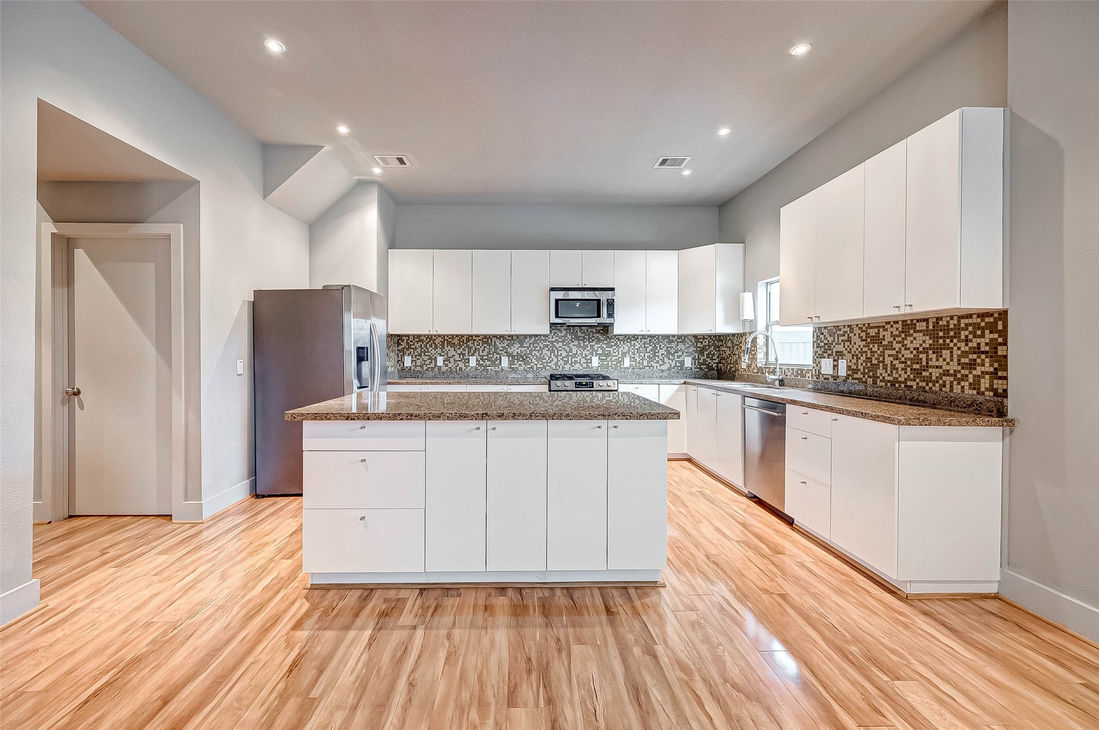 Spacious modern kitchen with sleek white cabinetry, granite countertops, and a stylish mosaic backsplash, complemented by stainless steel appliances and warm wood flooring.