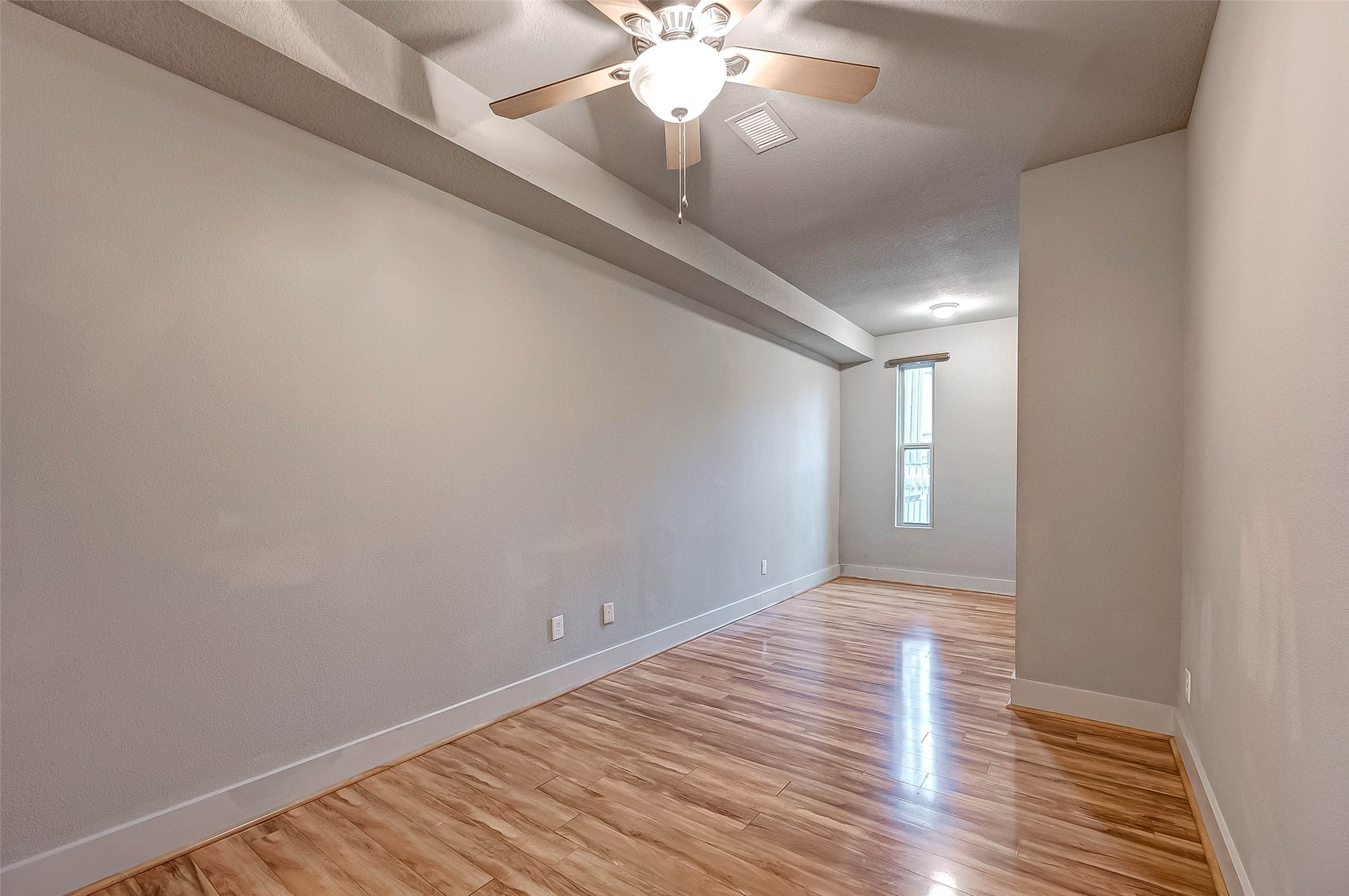 Spacious room on FIRST flood with sleek hardwood floors, neutral walls, and a ceiling fan for comfort, accented by natural light from a tall window.