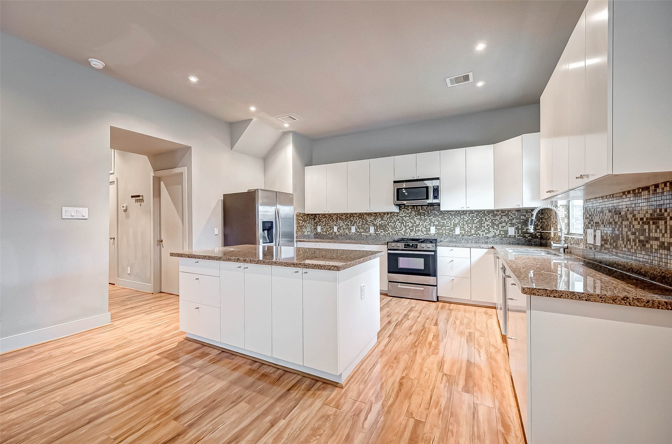 Spacious modern kitchen featuring sleek white cabinetry, granite countertops, and stainless steel appliances, complemented by a stylish mosaic tile backsplash and warm wood flooring.