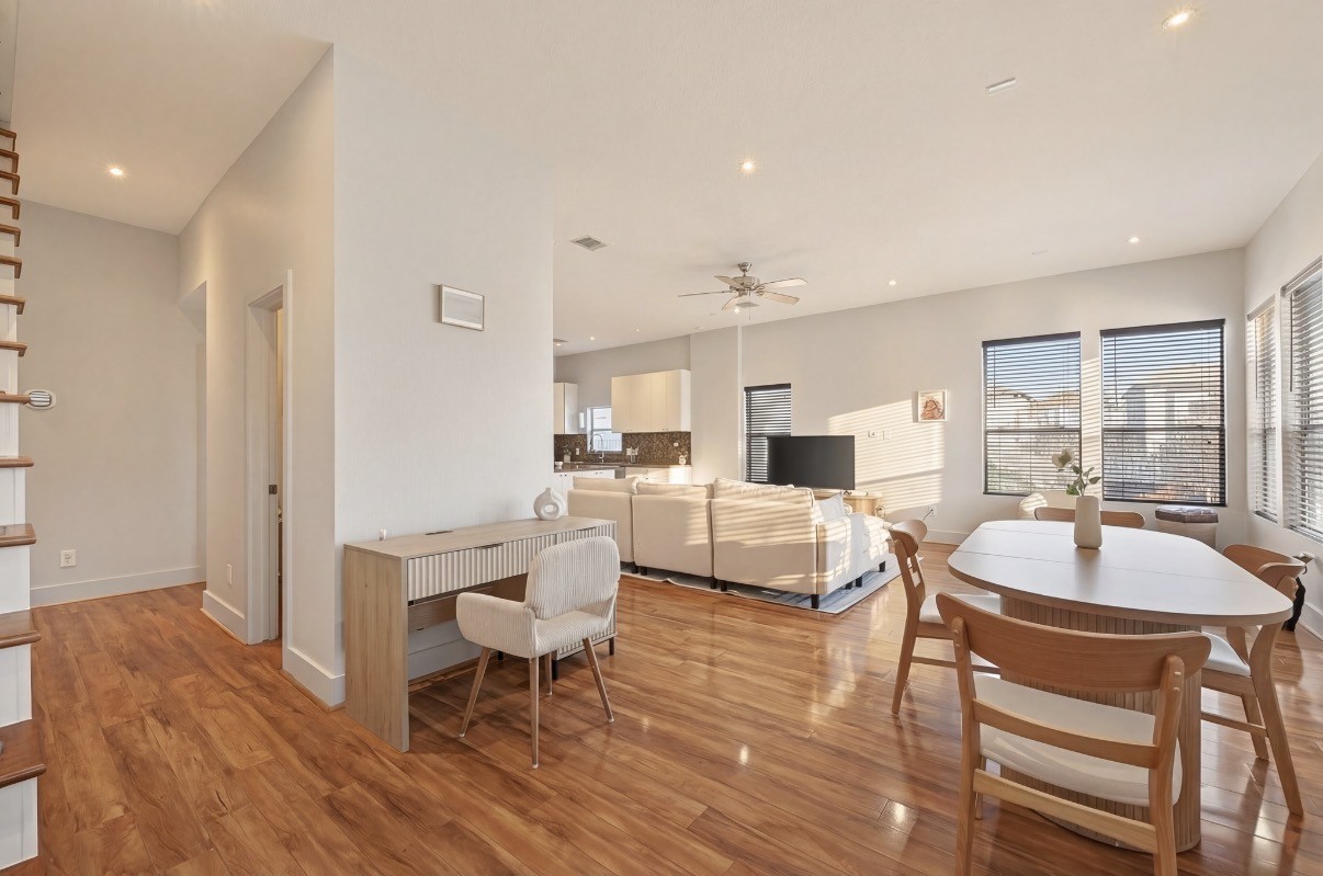 Bright and inviting open-concept living area featuring warm wood flooring, modern furnishings, ample natural light from large windows, and a cozy seating arrangement. Ideal for entertaining or relaxing.