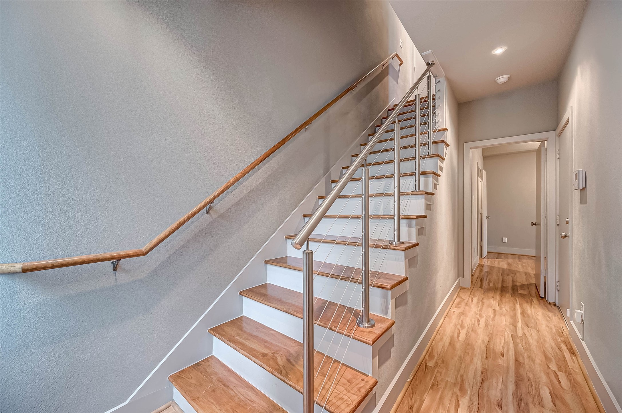 Modern staircase with sleek cable railing complements the bright, inviting hallway featuring warm wood flooring.