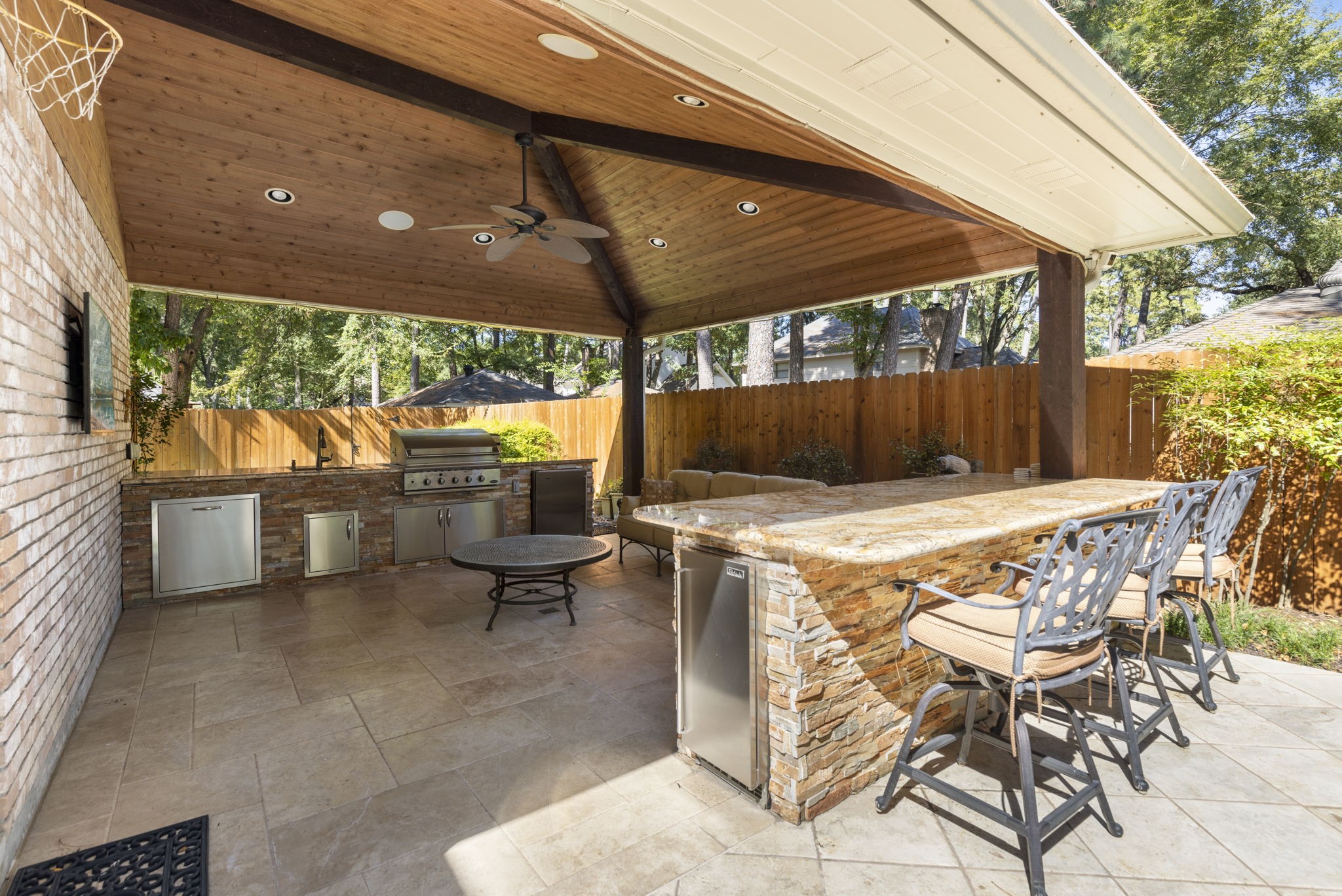 Beneath a wood ceiling, the covered outdoor kitchen features a built in grill, fridge, Perlic ice maker, and generous bar seating for relaxed poolside hosting.