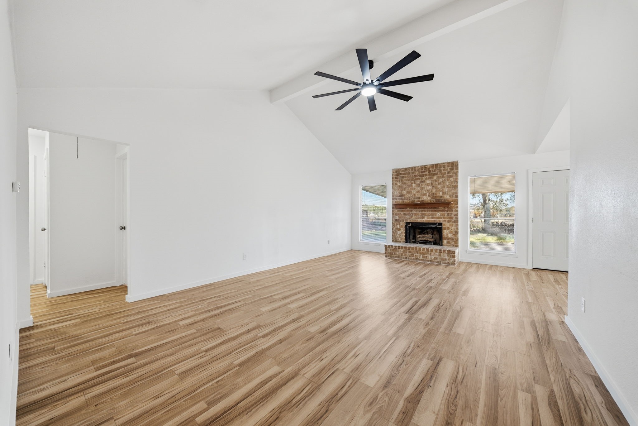 The same living room empty: vaulted ceiling, beam, brick gas fireplace and two yard-facing windows.