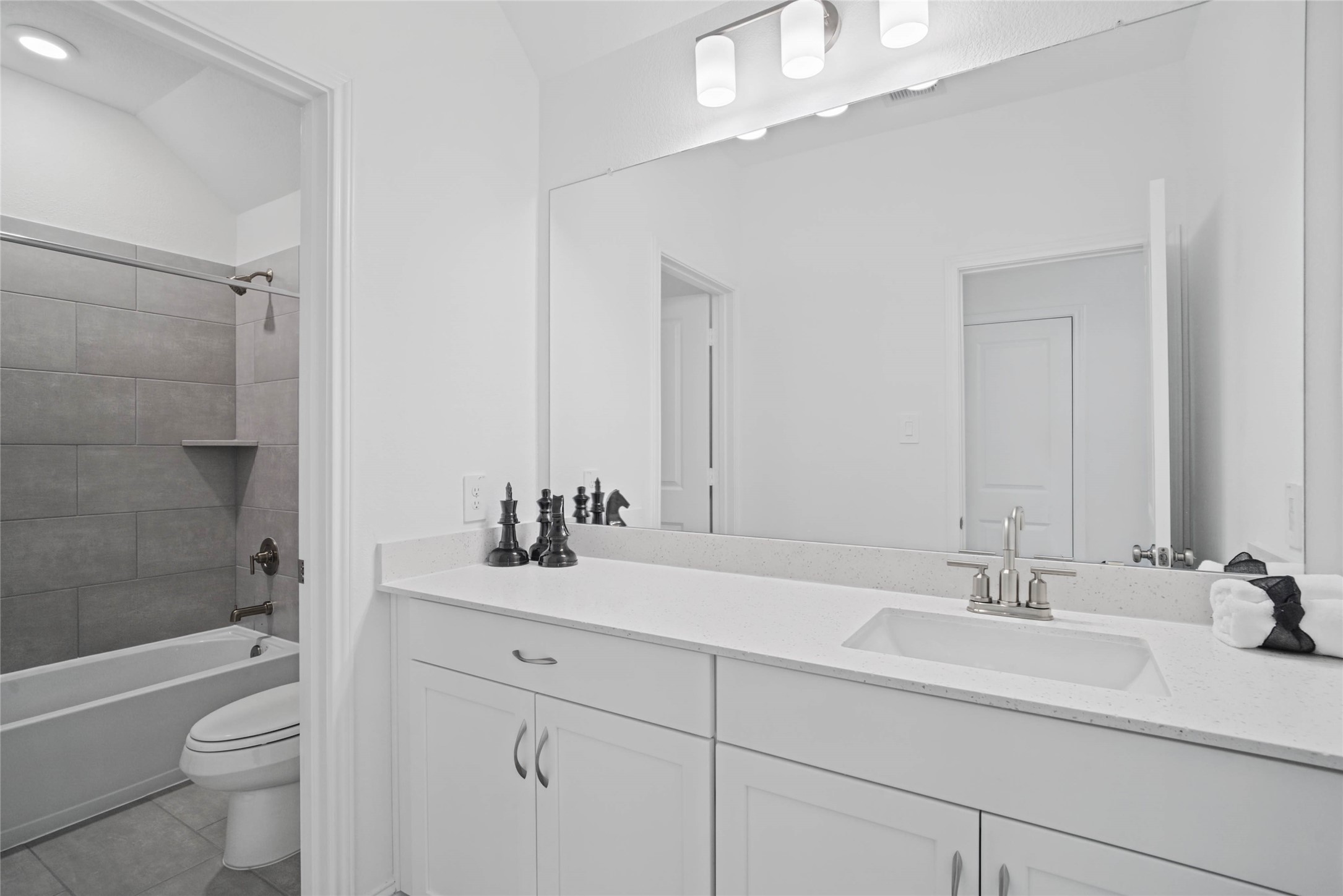 Secondary bathroom featuring an extended vanity cabinet with single bowl sink, brushed nickel hardware, mirror and vanity lighting, tub/shower combination with tile surround and tile flooring.