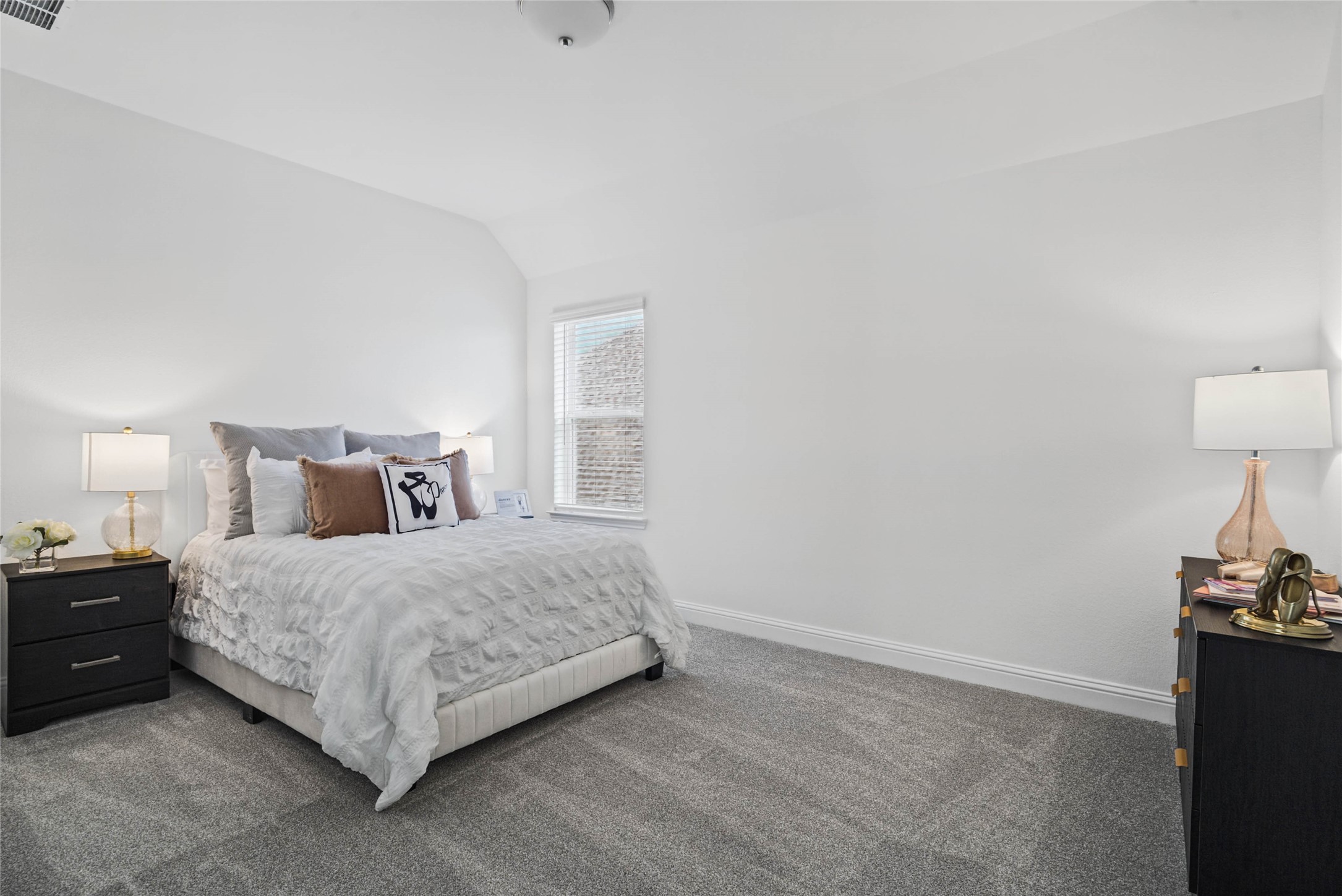 This inviting secondary bedroom combines comfort and modern elegance, featuring plush carpeting, a neutral color palette, and abundant natural light streaming through a large window with blinds.