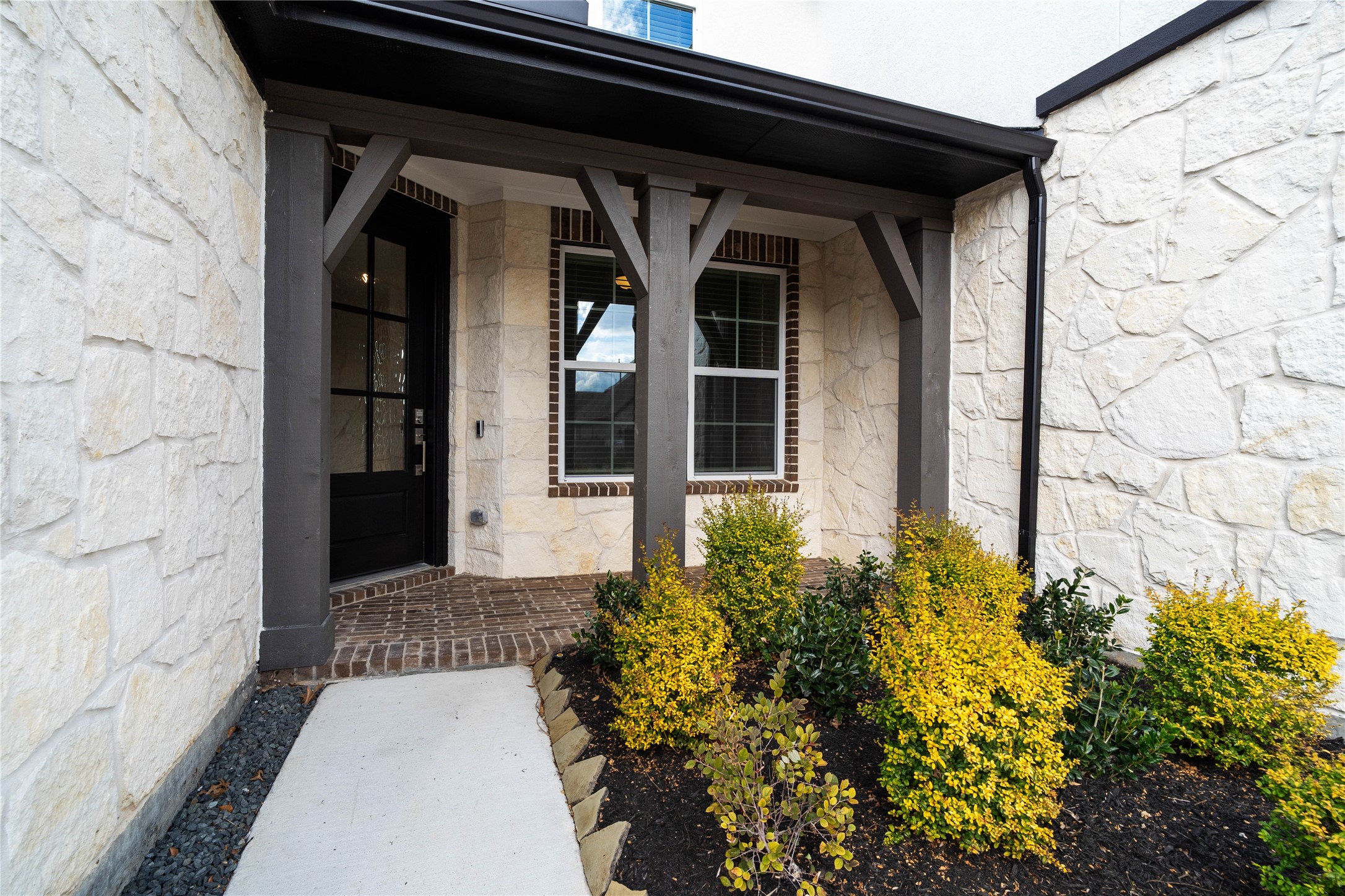 This covered front porch offers a grand first impression with its modern yet rustic aesthetic. The entrance features striking dark-stained timber beams and brackets that provide structural character against a beautiful white stone veneer faade.