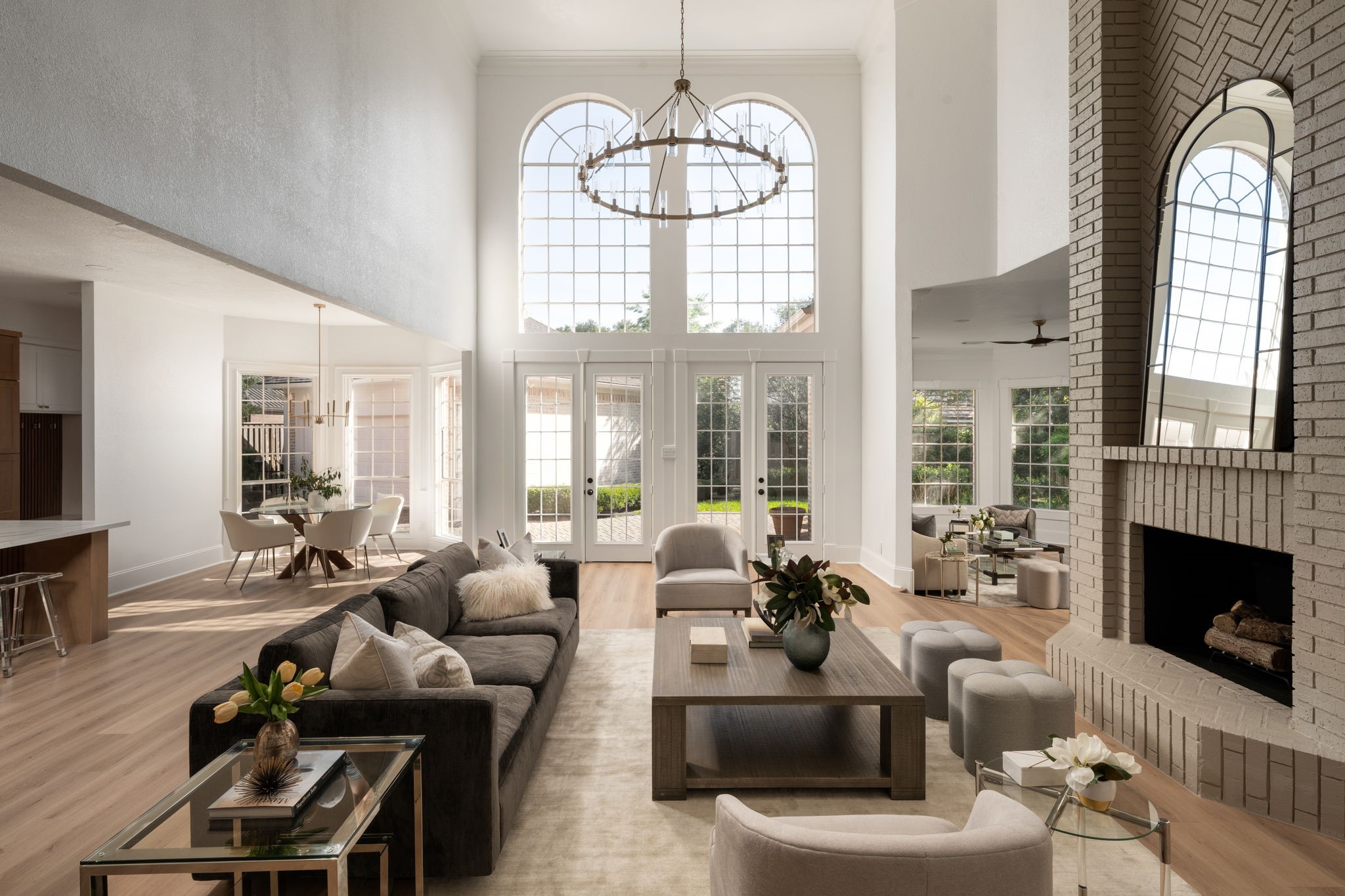 The main living area is defined by soaring double-height ceilings, expansive windows, and a layout designed for gathering and entertaining. Sunlight moves throughout the space during the day, highlighting the renovated flooring and clean architectural lines. A decorative fireplace feature anchors the room while maintaining connection to the kitchen and outdoor spaces.