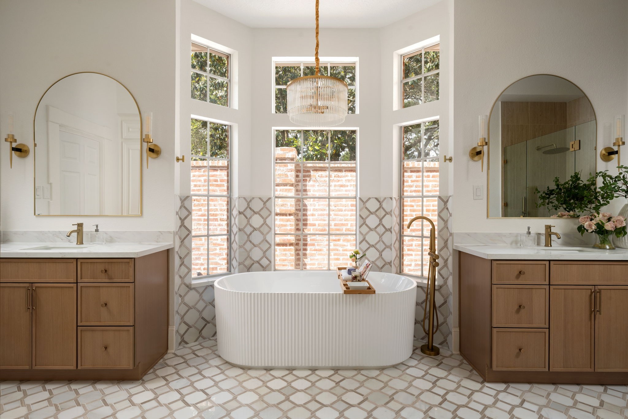 The primary bath features a freestanding soaking tub positioned beneath oversized windows and surrounded by custom marble flooring and tilework. Dual vanities, brass fixtures, and contemporary lighting contribute to the spaces spa-inspired design. Natural light fills the room throughout the day while maintaining privacy.