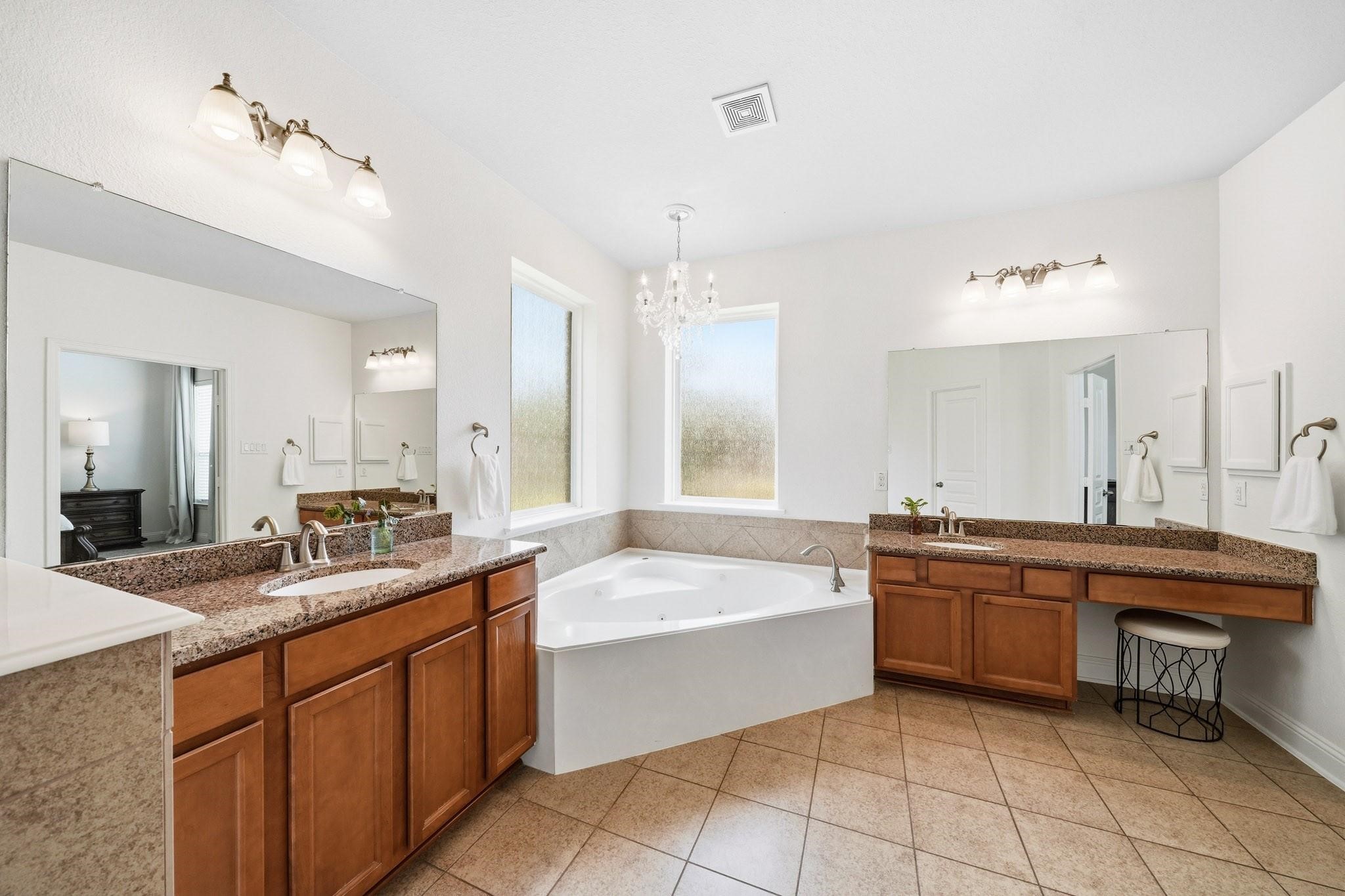 A six-foot jetted tub serves as the centerpiece of the bathroom, perfectly positioned beneath dual rain-glass picture windows and illuminated by a sparkling chandelier, creating a spa-inspired retreat.