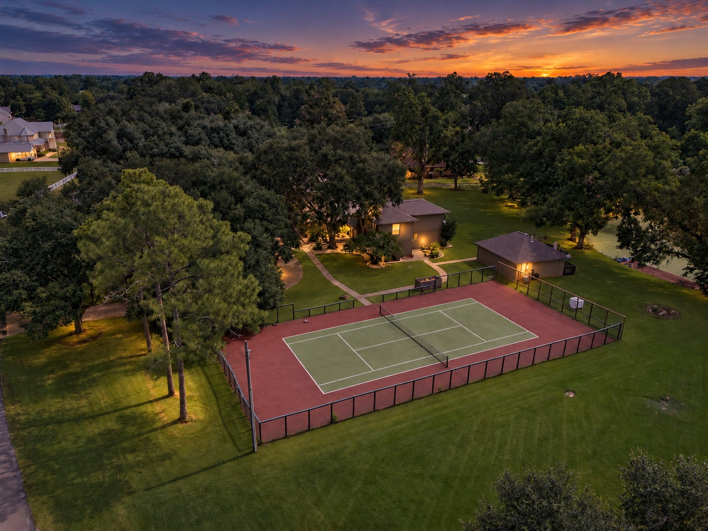 PRIVATE TENNIS COURT SURROUNDED BY EXPANSIVE LAWN AND MATURE TREES, CREATING A SPACIOUS OUTDOOR RECREATION AREA.