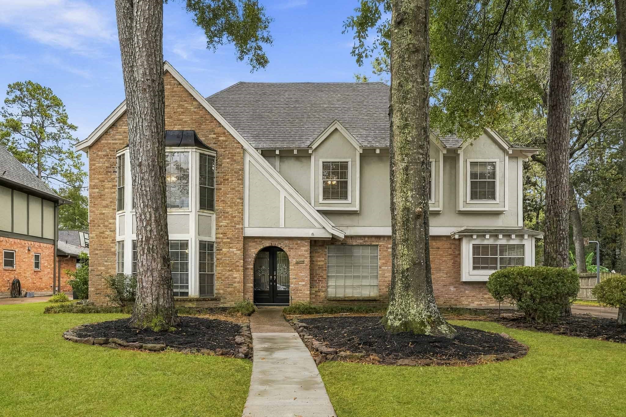 Charming two-story home with a classic brick and stucco exterior, featuring a welcoming archway entrance and large bay windows. Nestled among mature trees, this property boasts a well-manicured lawn and a serene suburban setting.