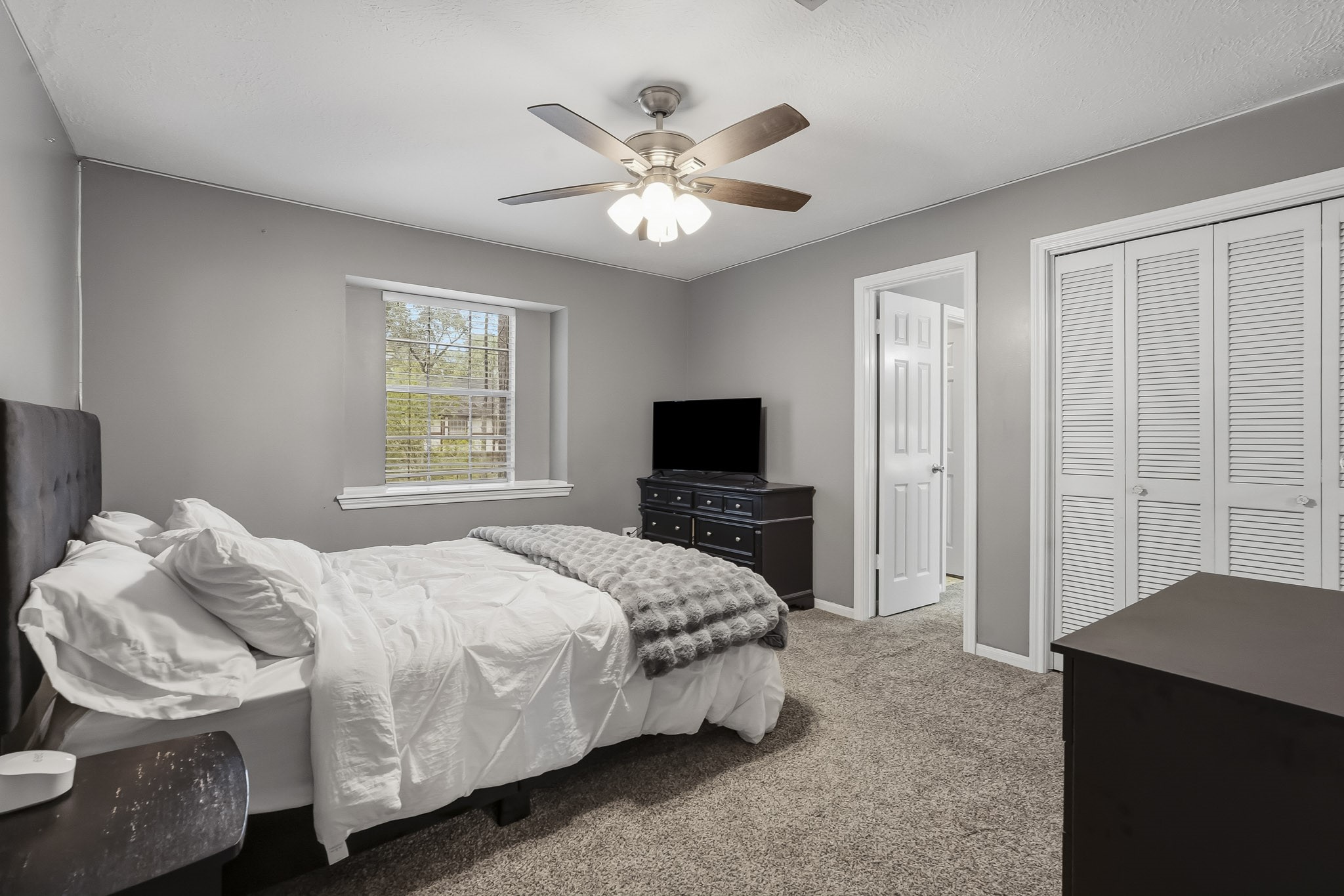 Spacious third bedroom with soothing gray walls, plush carpeting, and a ceiling fan. Features a large window for natural light, ample closet space, and an en-suite bathroom.