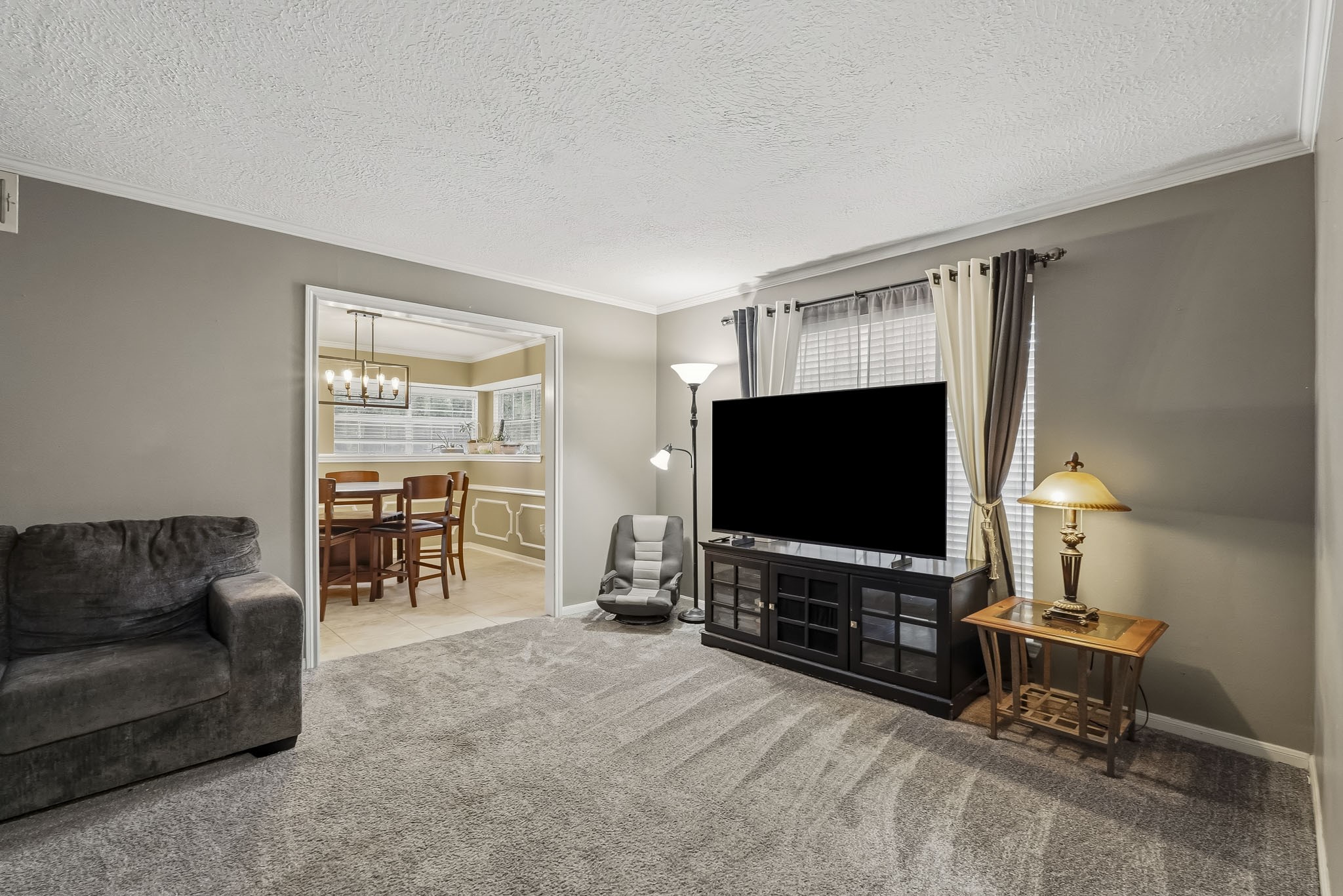 Cozy flex room or formal living with plush carpeting and a large window for natural light, seamlessly connected to a dining area with elegant lighting.