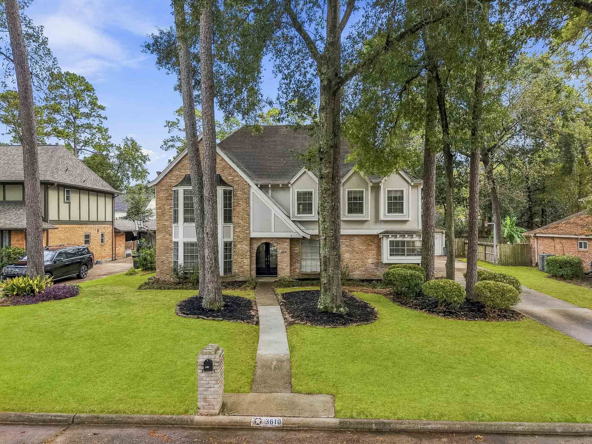 Charming tudor style two-story brick home with elegant gables, nestled among towering trees and lush greenery, featuring a welcoming pathway and well-maintained landscaping.
