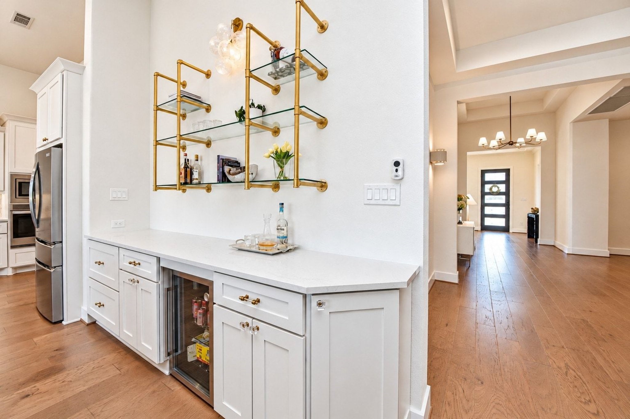 Custom open shelving unit with gold accents features a built-in beverage refrigerator. The adjoining area flows seamlessly into a bright hallway with stylish lighting and wood flooring.