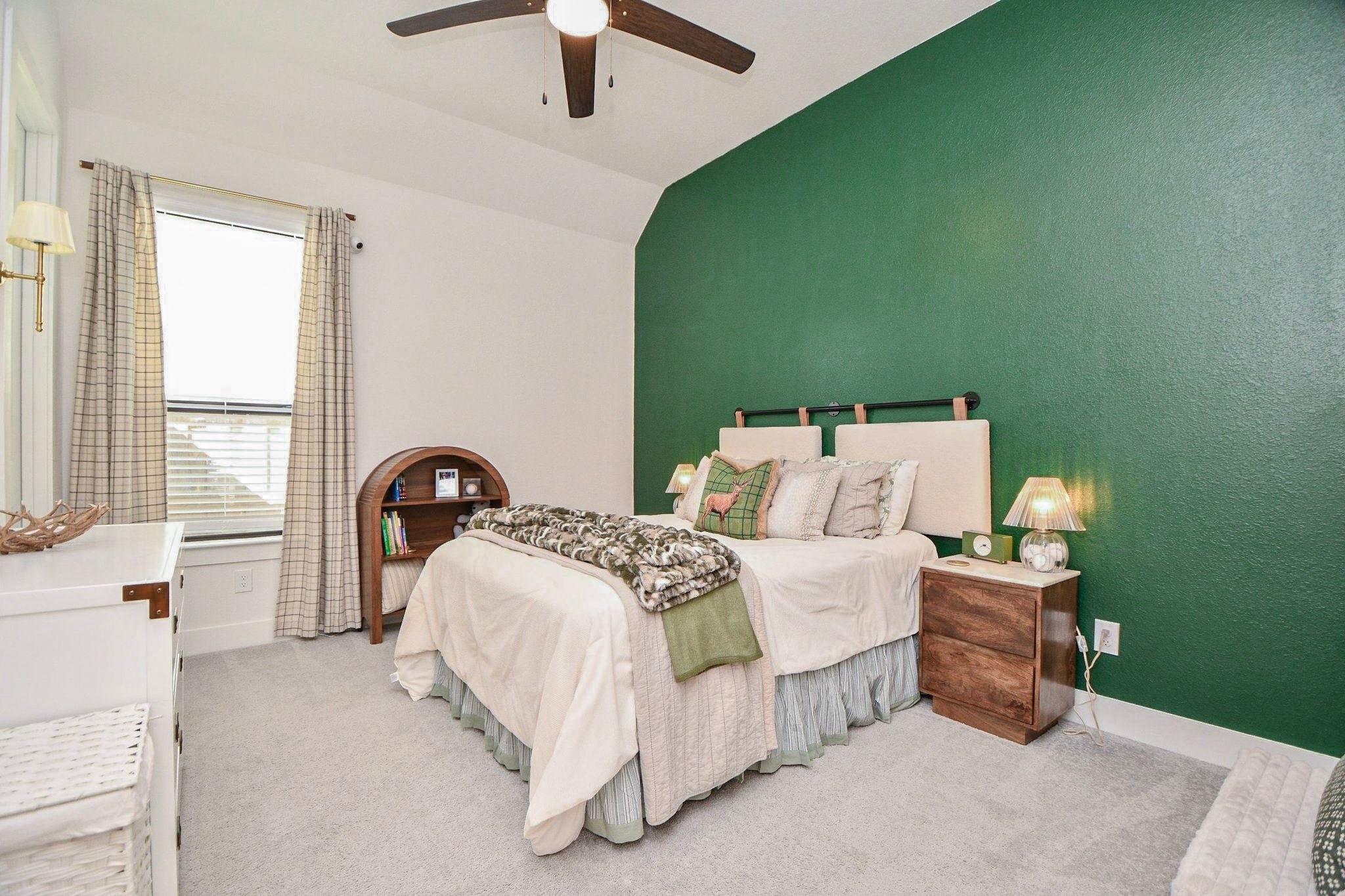 Cozy bedroom with a striking green accent wall, plush carpeting, and a ceiling fan. Natural light filters through the window, complementing the warm, inviting decor.