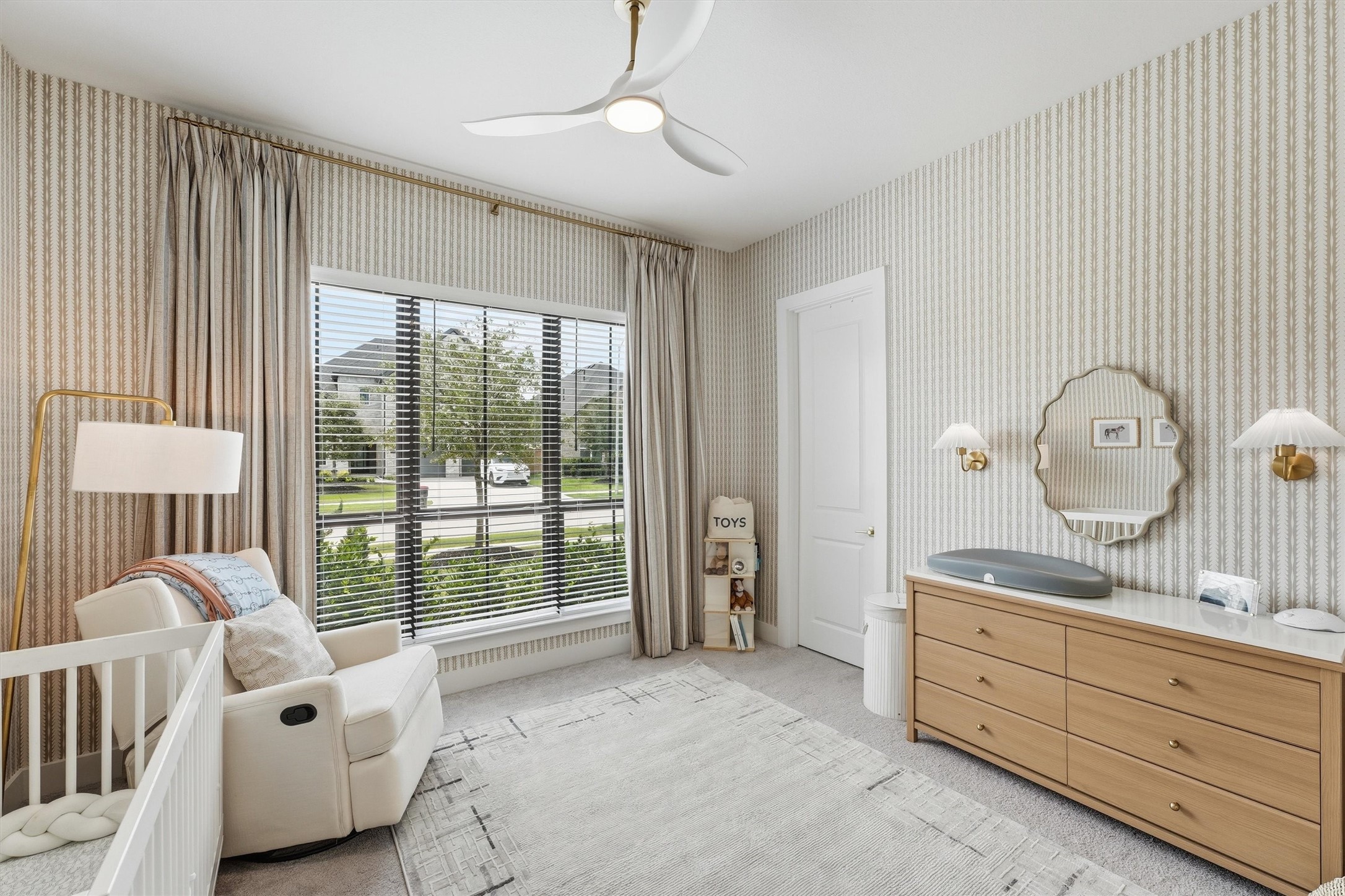 A delightful secondary bedroom or nursery bathed in natural light, featuring soft patterned wallpaper, elegant window treatments, modern lighting, and plush carpeting, creating a warm and inviting space full of charm.