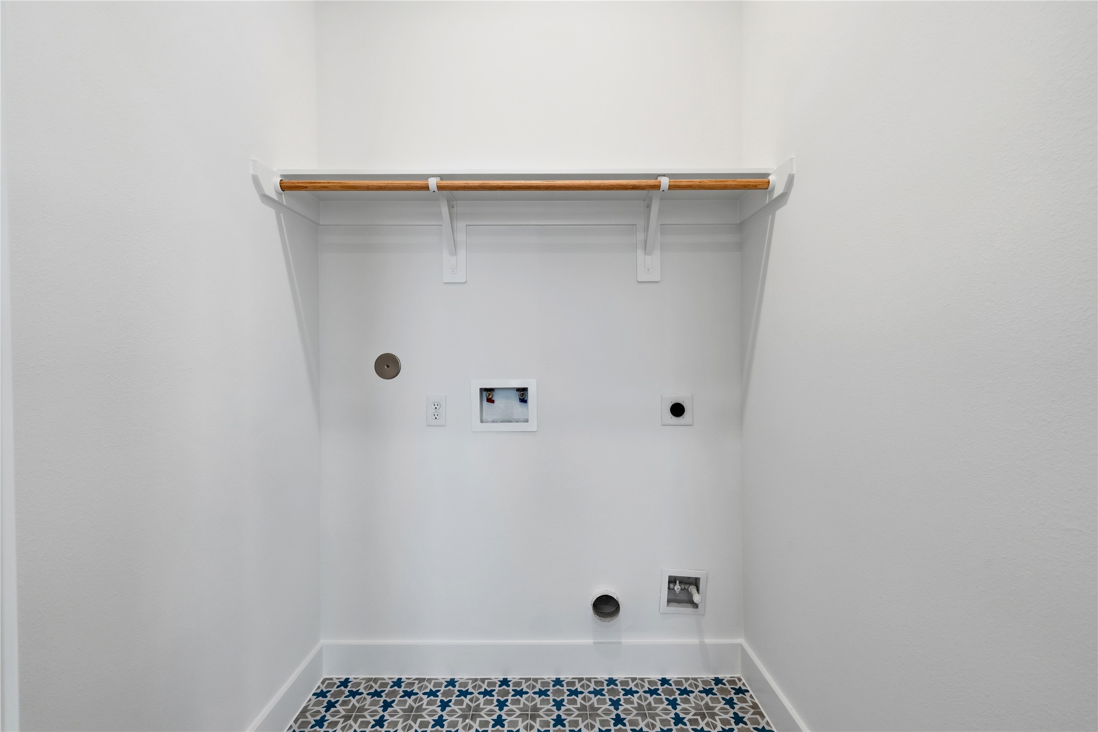 A dedicated laundry alcove featuring patterned Mediterranean-style floor tiles and a natural wood hanging rod. The minimalist white environment includes integrated plumbing and electrical connections for a streamlined look.