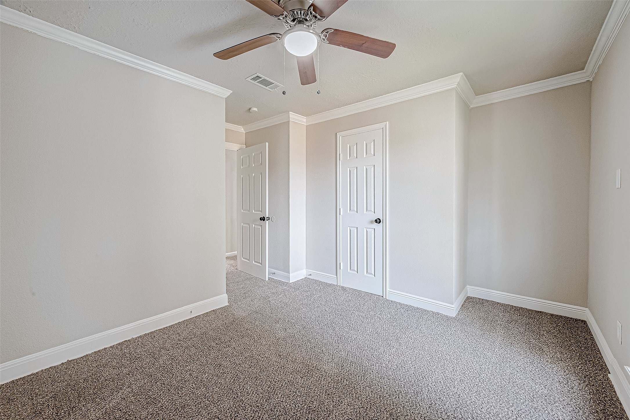 Bright and airy room with neutral tones, featuring carpet flooring, a ceiling fan, two doors, and elegant crown molding.
