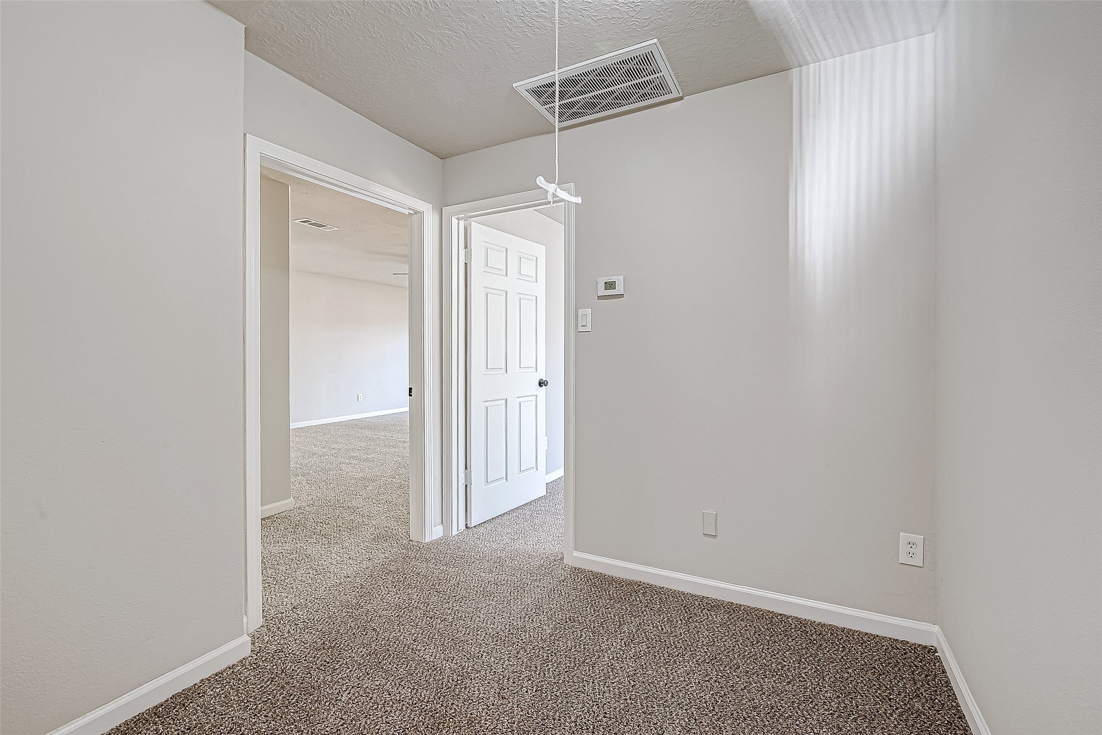A small, carpeted room with neutral walls, leading to a larger space through an open doorway.