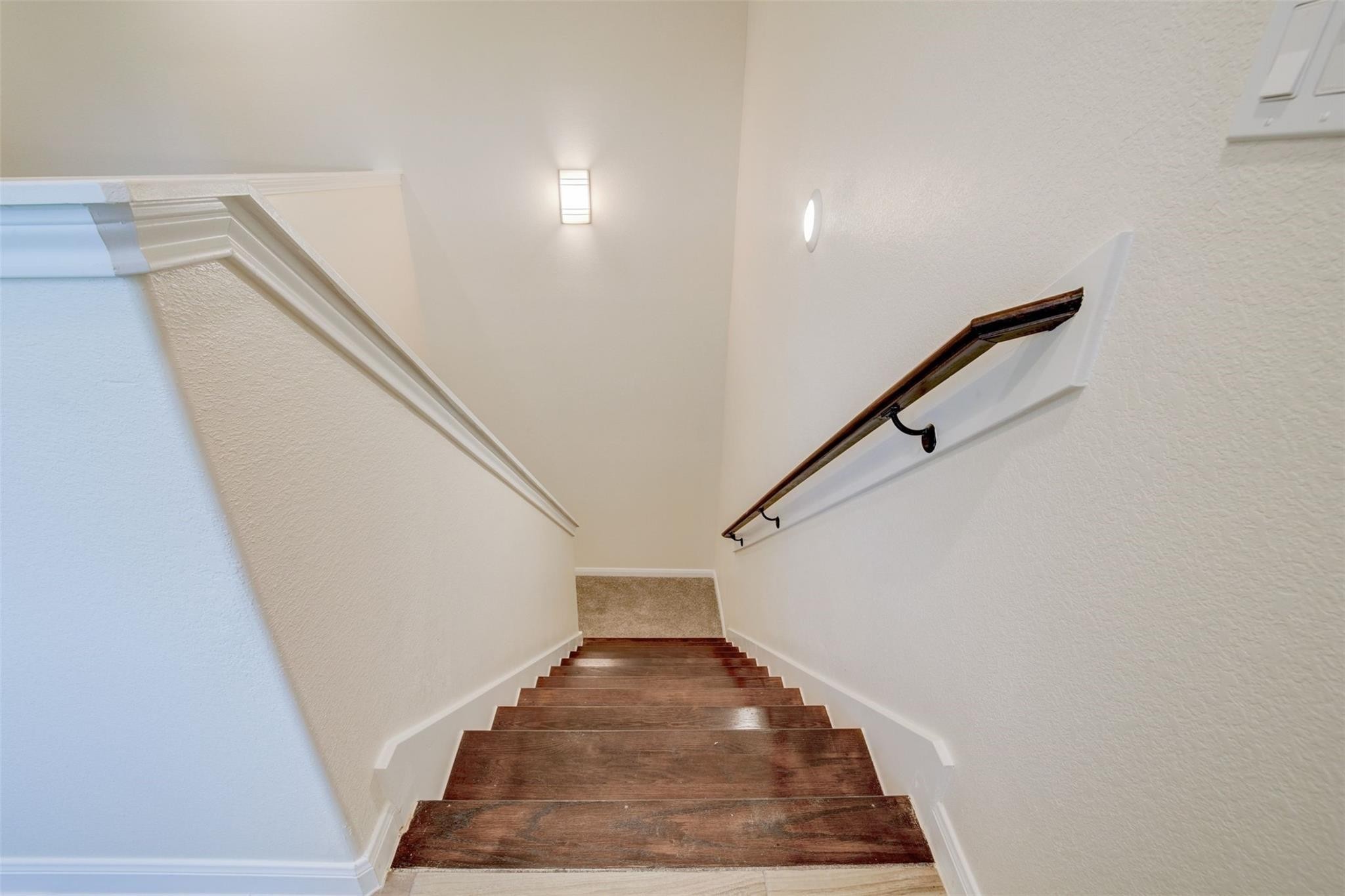 A well-lit staircase with wooden steps and a sleek handrail on the right side, leading to upstairs level. The walls are painted in a light, neutral color, enhancing the bright and open feel.