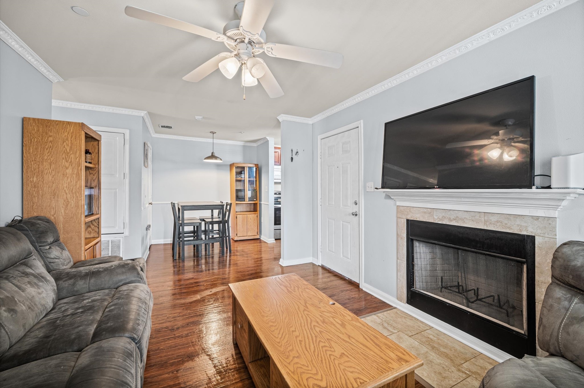 Cozy living space with a ceiling fan, featuring a fireplace. The open layout flows into a dining area and kitchen, accented by wood flooring and built-in shelving.