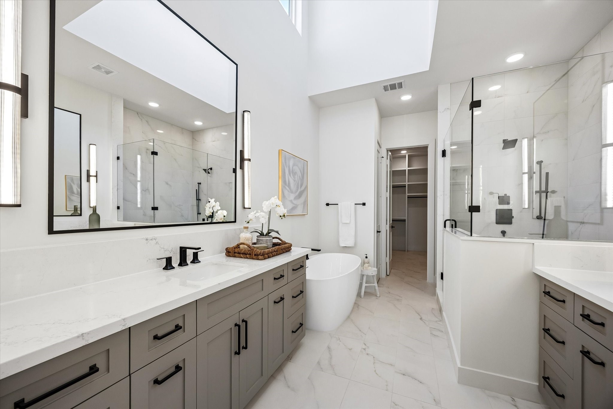 Indulge in a spa-like experience in the primary bath, featuring soaring ceilings, ample natural light from a large window, and double custom-built vanities with Kohler undermount sinks. Visual Comfort sconces frame the mirrors, and sleek porcelain tile floors create a chic, modern look. Straight ahead the door opens to a huge walk-in closet with his and hers dressers and hidden storage that completes the ultimate retreat.