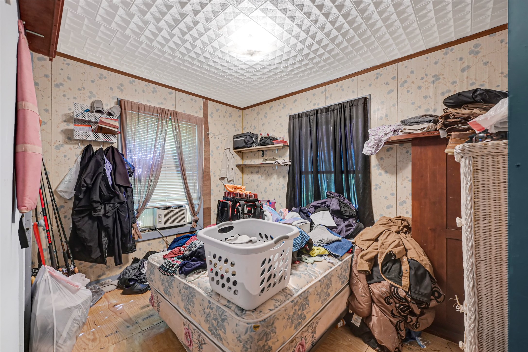 Cozy bedroom with potential, featuring textured ceilings and floral wallpaper. Ample natural light filters through dual windows, complemented by practical storage solutions. Ideal for personalization and creativity.