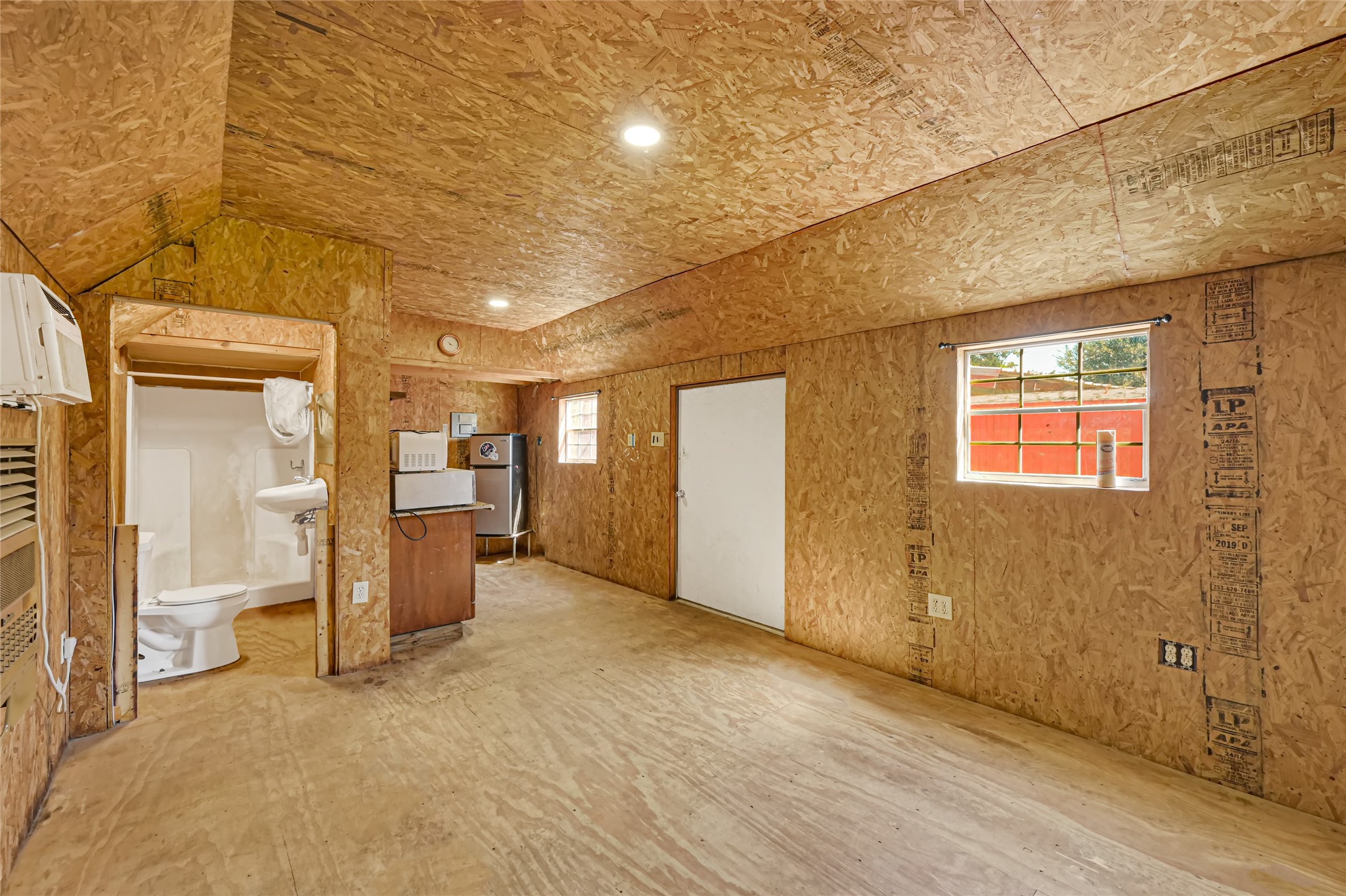 Spacious room with plywood walls featuring a small kitchenette and a convenient half-bath, offering a blank slate for customization.
