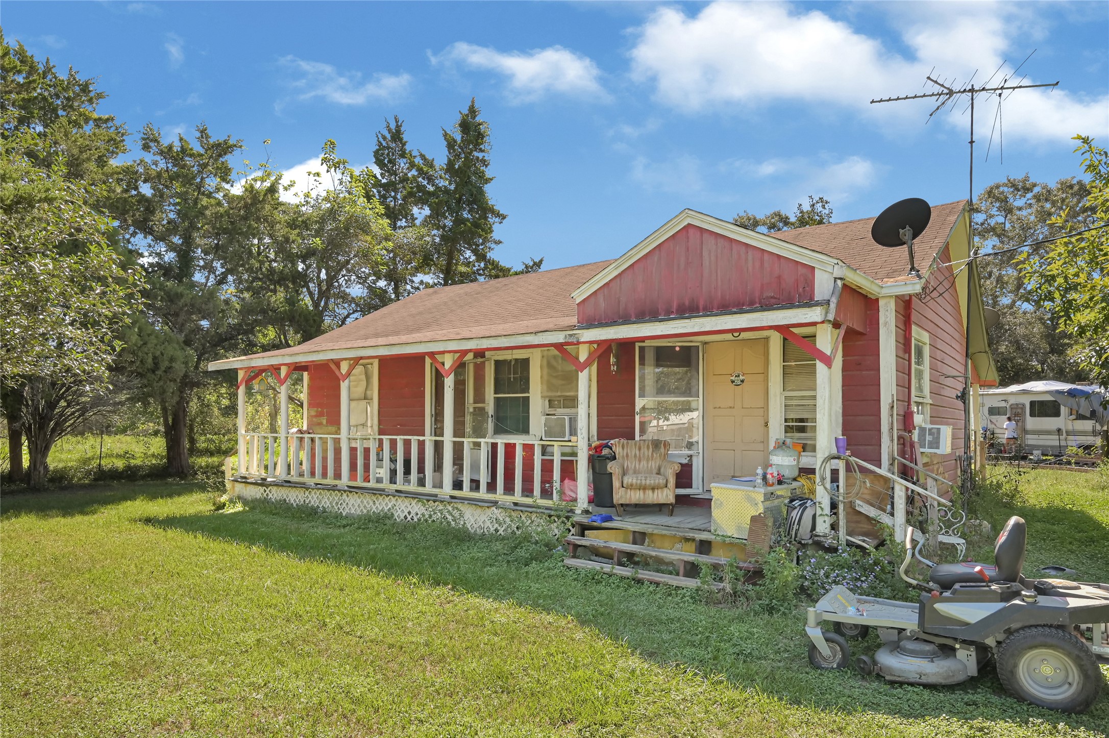 Charming red cottage with a cozy front porch, surrounded by lush greenery and mature trees, offering a peaceful, rural ambiance.