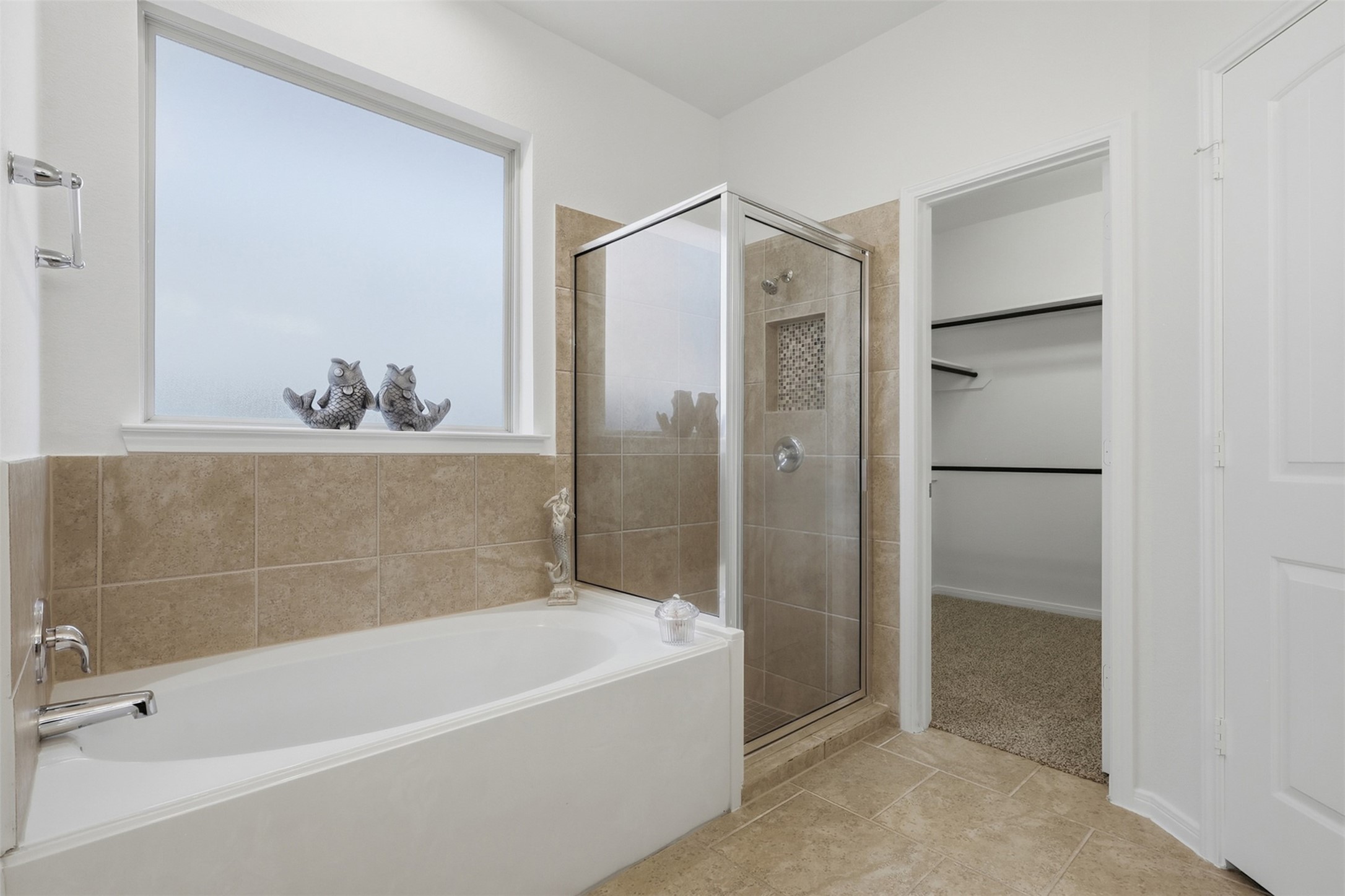 Relax, Refresh & Enjoy! This inviting primary bath showcases a beautiful soaking tub positioned beneath a large window, a separate glass-enclosed shower, and a spacious walk-in closet just beyond. The warm tile finishes and abundant natural light give the room a bright, comfortable feel. A wonderful private retreat within this lovely primary suite!