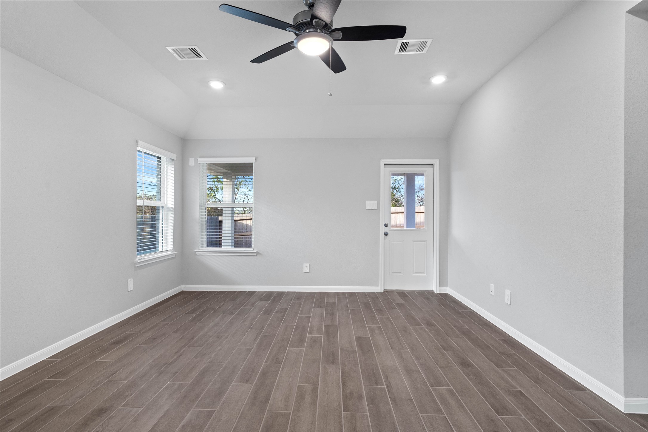 A bright and airy open-concept space featuring tile flooring, sloped ceilings with recessed lighting and ceiling fan, abundant natural light from large windows (fitted with blinds), and an access door to the back covered patio.