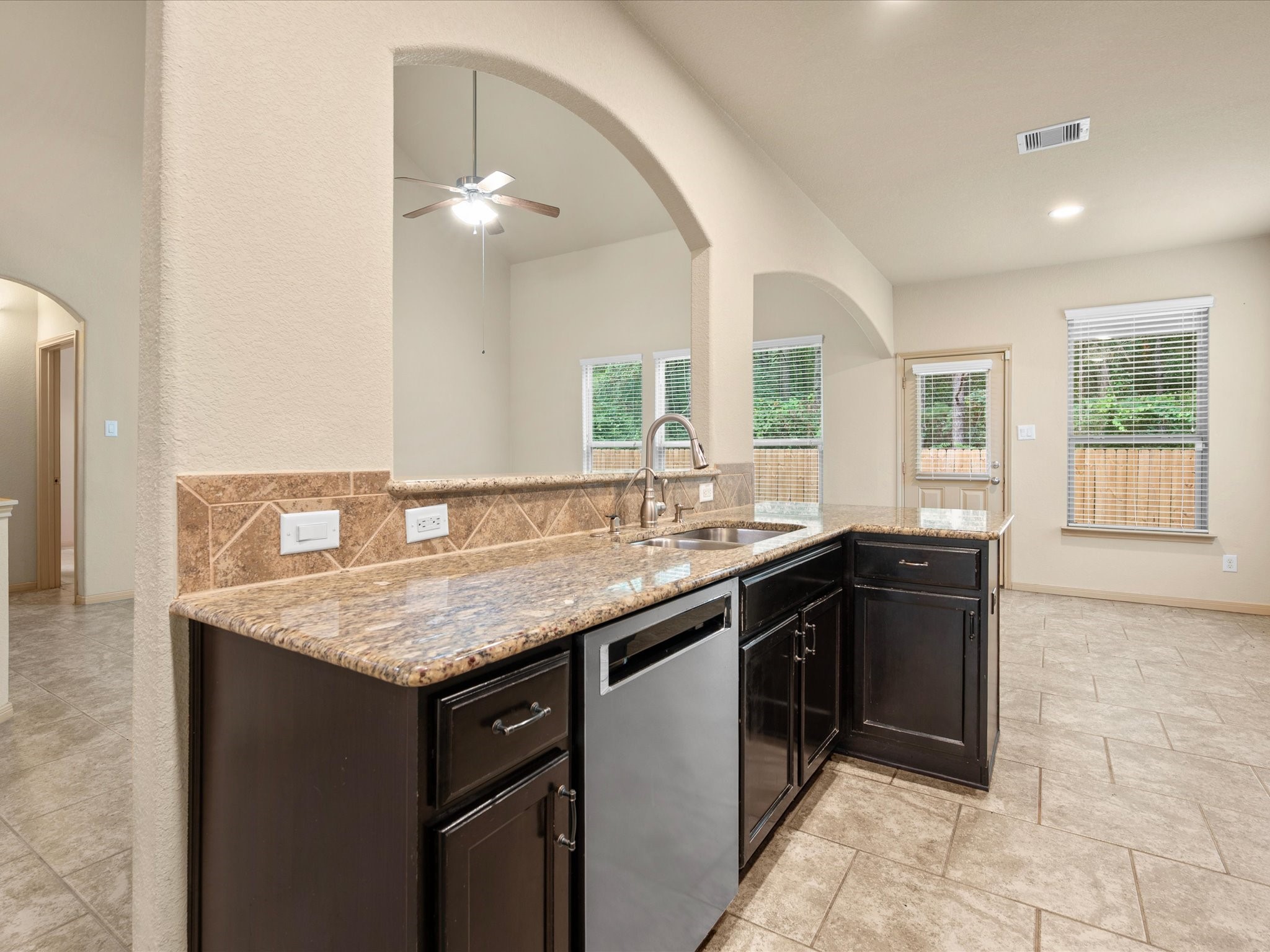 Kitchen viewpoint showcasing granite countertops, dark rich cabinetry, and a stainless steel dishwasher. An arched pass-through cutout above the double sink keeps you connected to the main living room while framing views of the adjacent light-filled breakfast nook and backyard entrance.