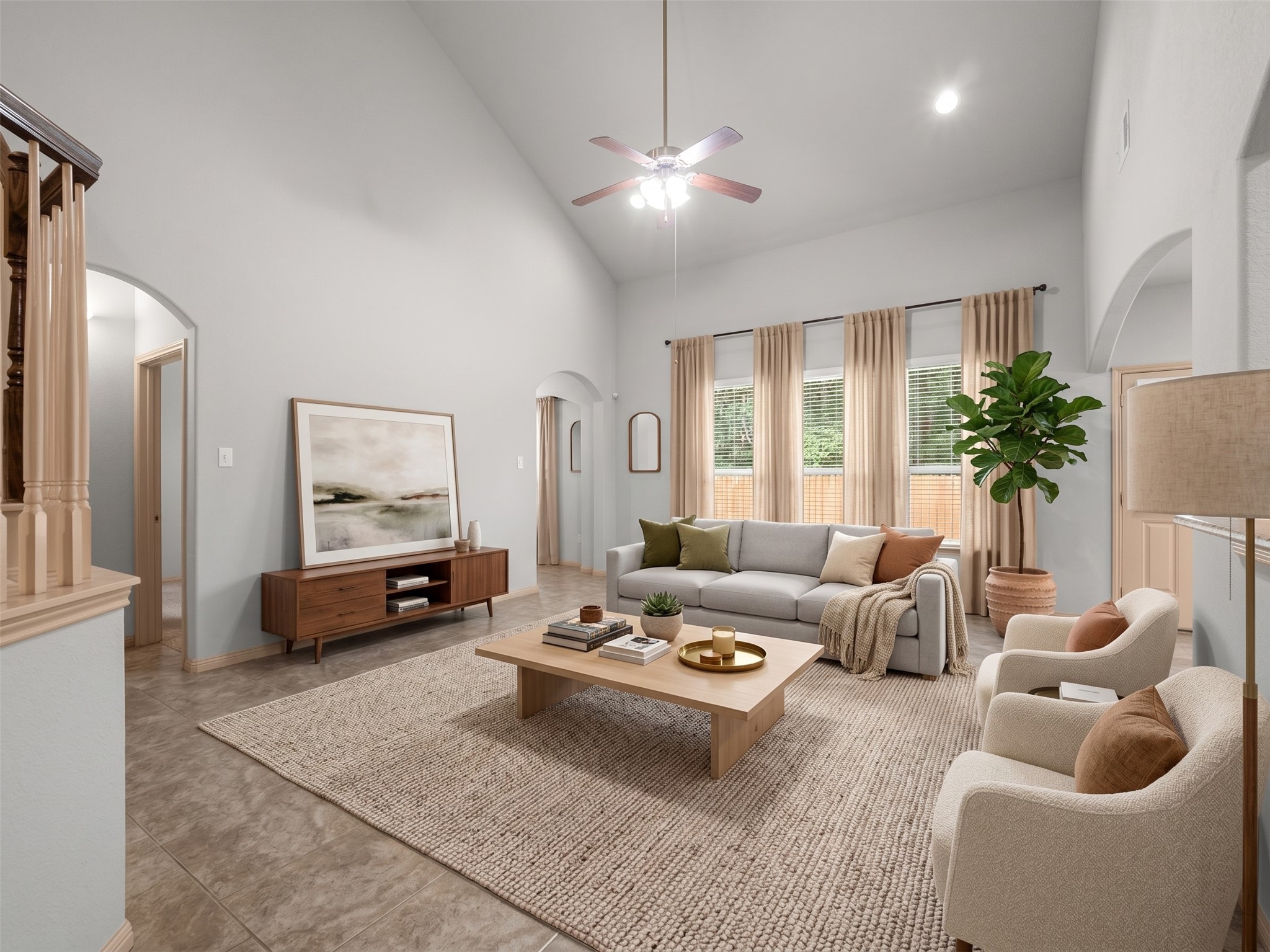Virtually staged living room showcasing the home's impressive vaulted ceilings and spacious floor plan. Features a modern light grey sofa, plush accent armchairs, a large neutral area rug over the tile flooring, and floor-to-ceiling drapery framing the backyard windows.