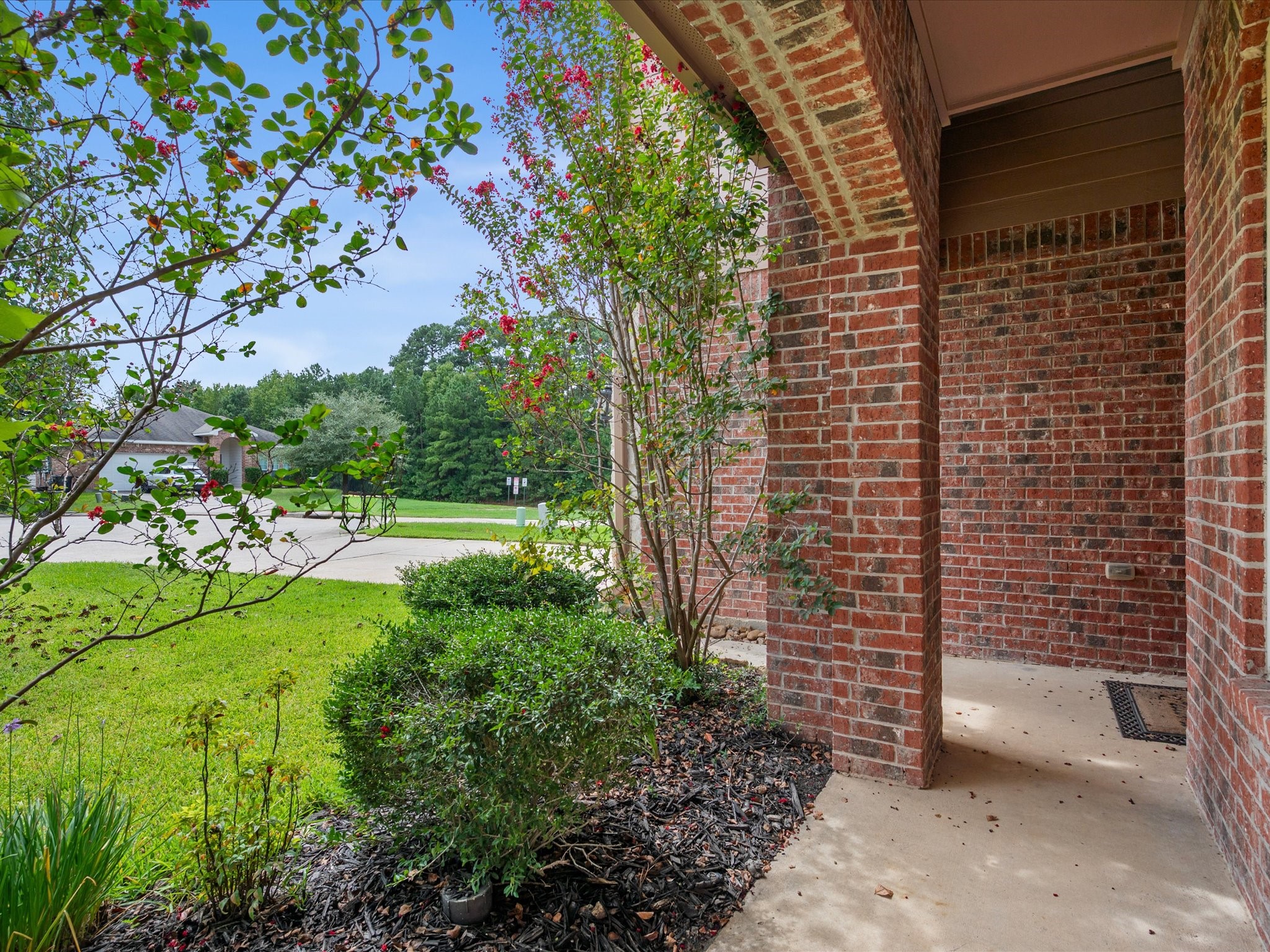 View looking out from the covered front porch framed by a classic brick archway. Highlights the peaceful neighborhood surroundings, manicured front garden beds with dark mulch, and lush flowering landscaping.