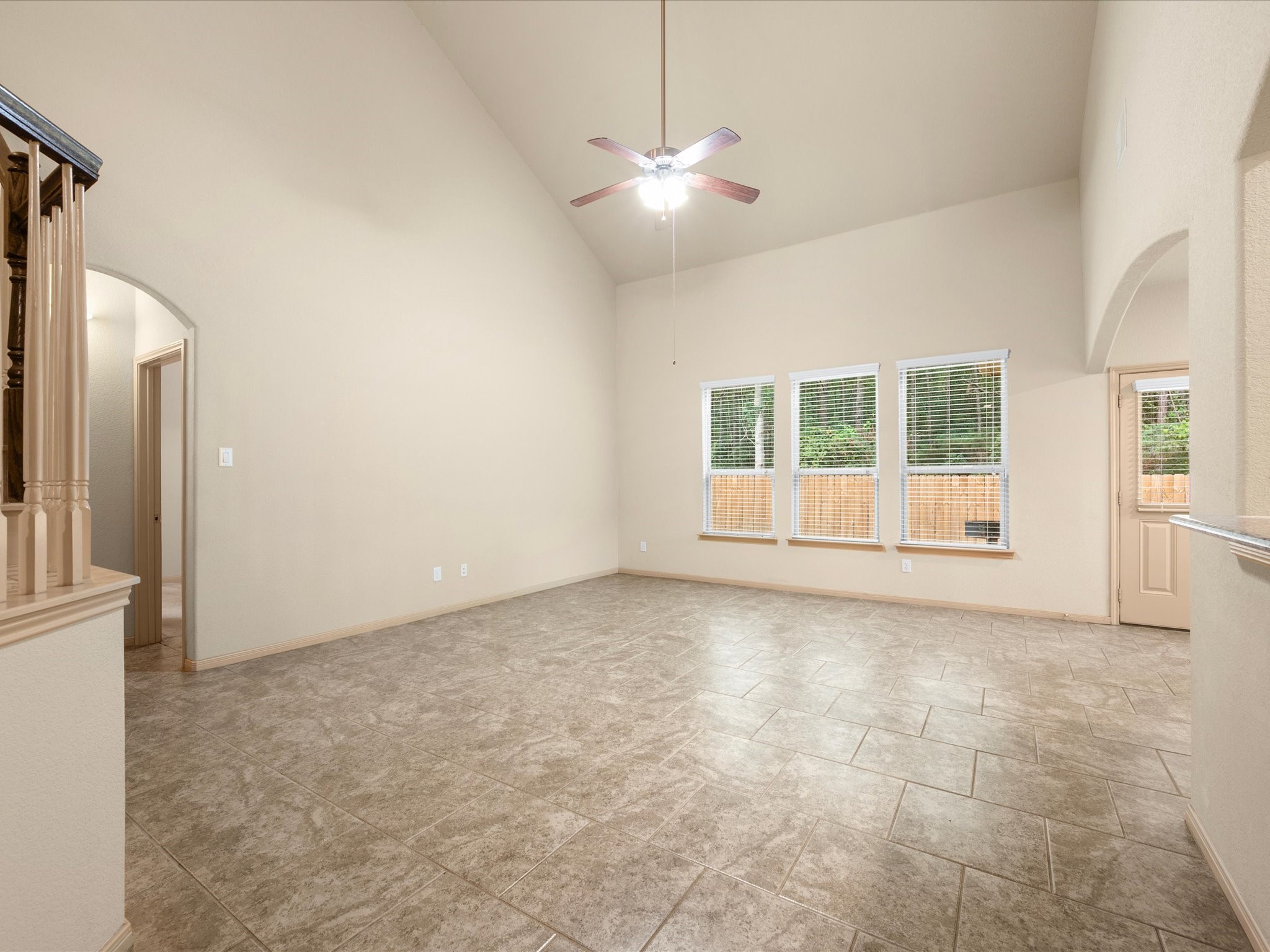 Spacious family room featuring soaring vaulted ceilings, a central ceiling fan, and durable tile flooring throughout. A wall of large windows floods the space with natural light while offering views of the private backyard.