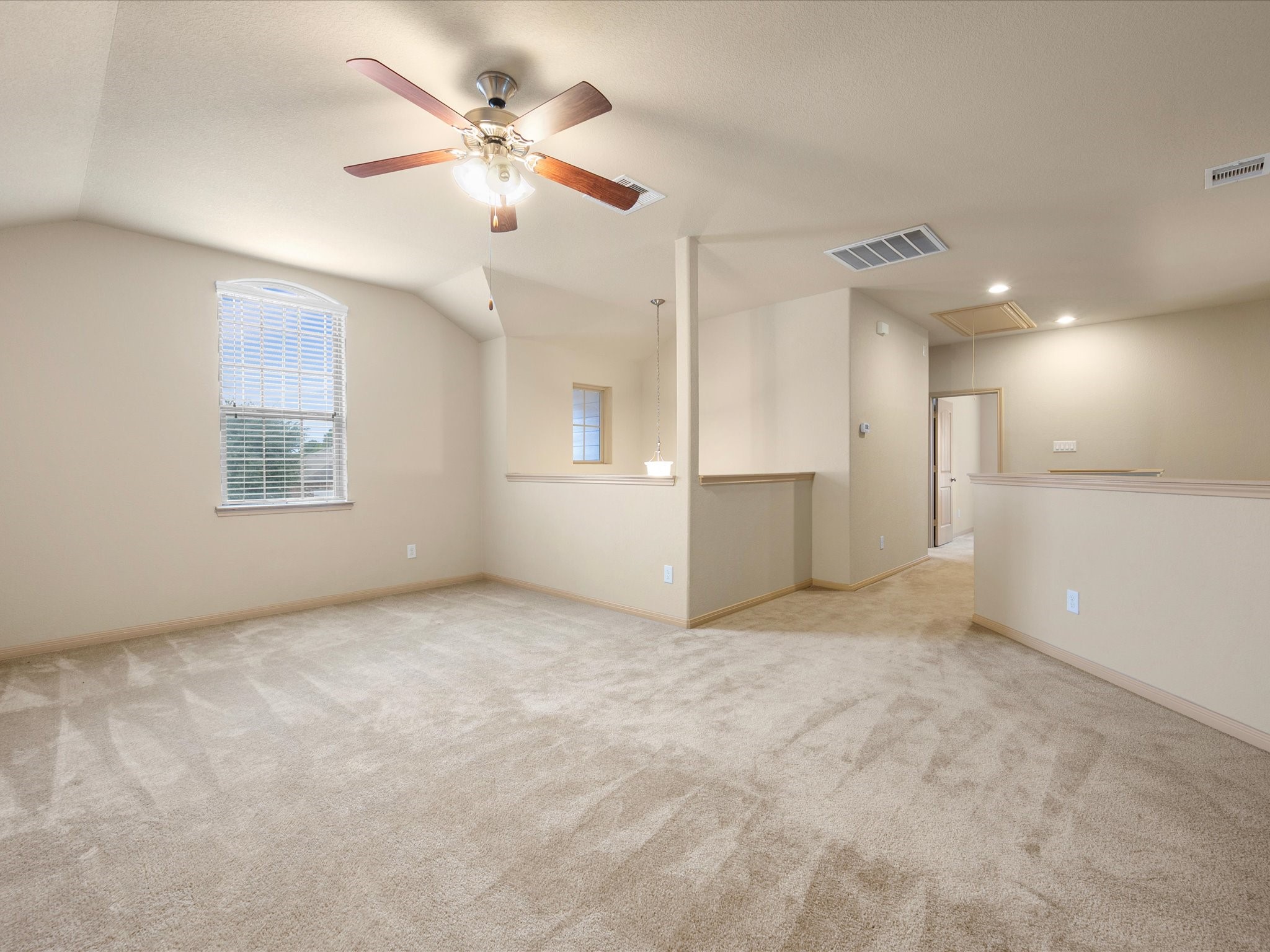 Expansive upper-level loft offering vaulted ceilings, a central ceiling fan, an arched window for natural light, plush carpet flooring, and partial wall dividers overlooking the stairs.