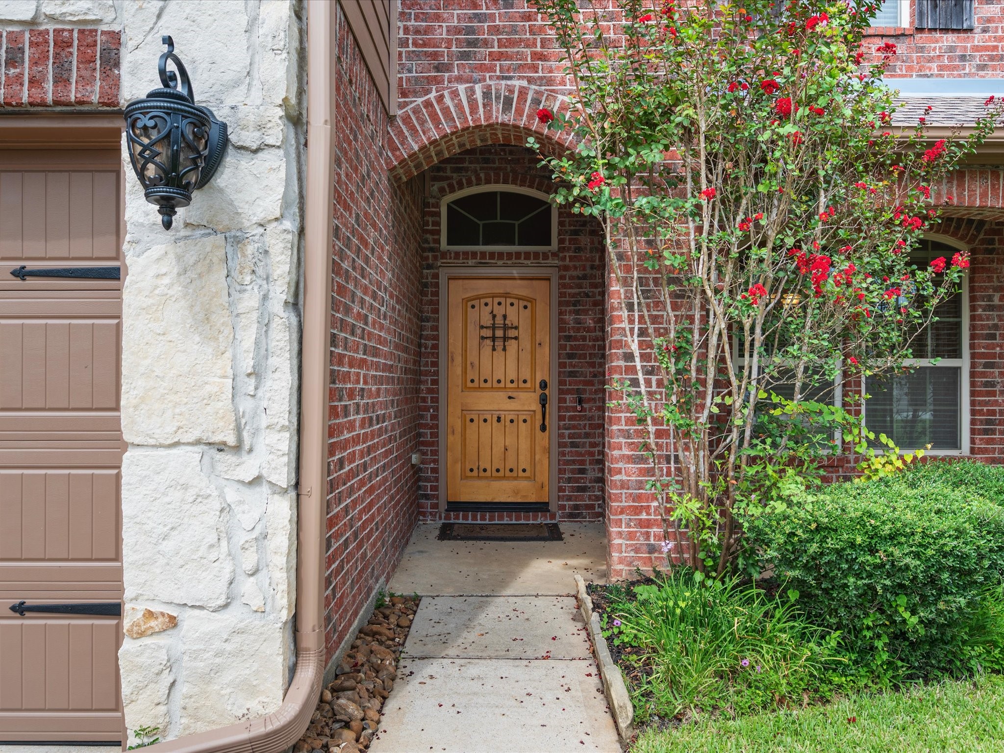 Welcoming covered entryway featuring a rustic wooden front door with ornate iron grill work and a transom window above. Surrounded by rich brick masonry, stone accents, classic exterior lantern lighting, and lush flowering landscaping.