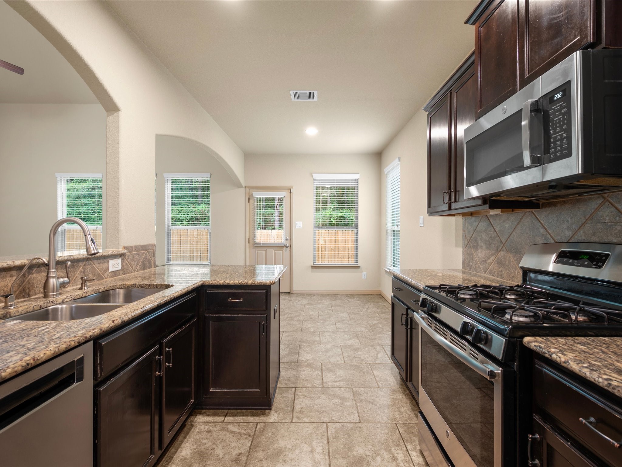 Kitchen view looking into the bright breakfast area, highlighting granite countertops, dark espresso cabinets, stainless steel gas appliances, and an arched pass-through to the family room.