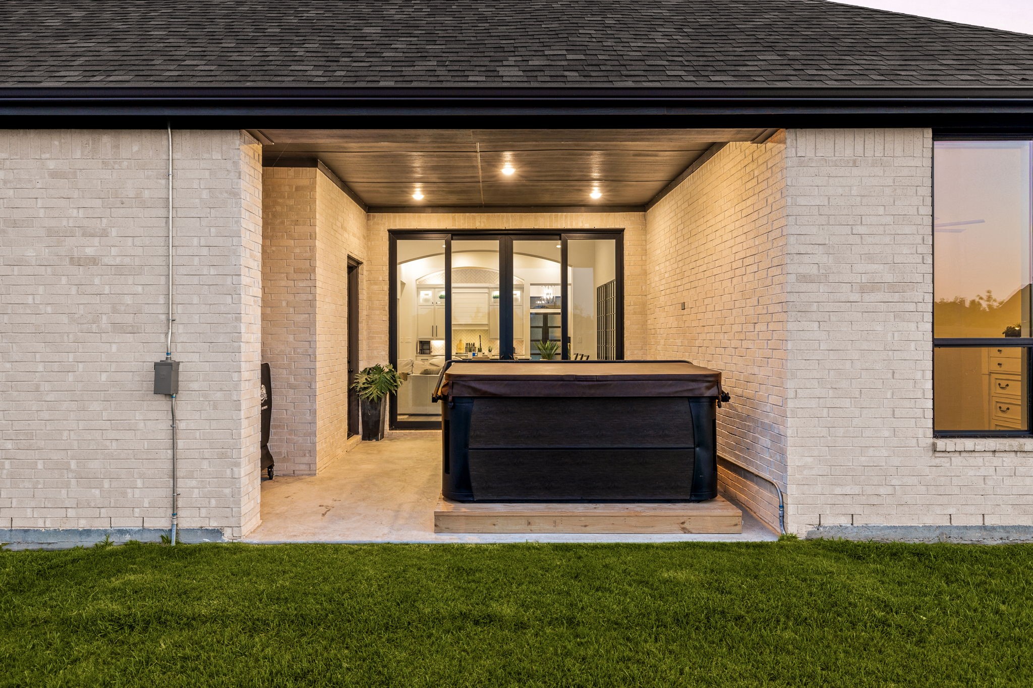 An inviting outdoor living space. This covered backyard patio features clean light brick columns, dark gutters, recessed ceiling lighting, a concrete floor, and glass slider doors that create a seamless indoor-outdoor transition.