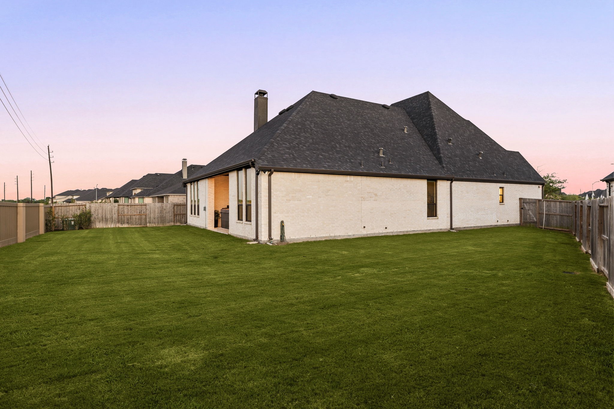 A pristine, low-maintenance exterior profile. This side view showcases the home's crisp light brick facade, clean rooflines, a continuous wooden privacy fence, and an expansive grassy lawn offering plenty of space for outdoor recreation.