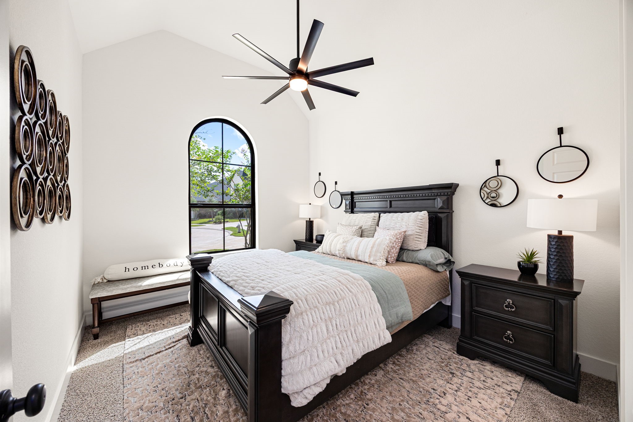 A charming and bright guest suite. A beautiful black-trimmed arched window anchors this inviting bedroom, highlighting plush neutral carpeting, soaring vaulted ceilings, a modern multi-blade ceiling fan, and clean matte black door hardware.