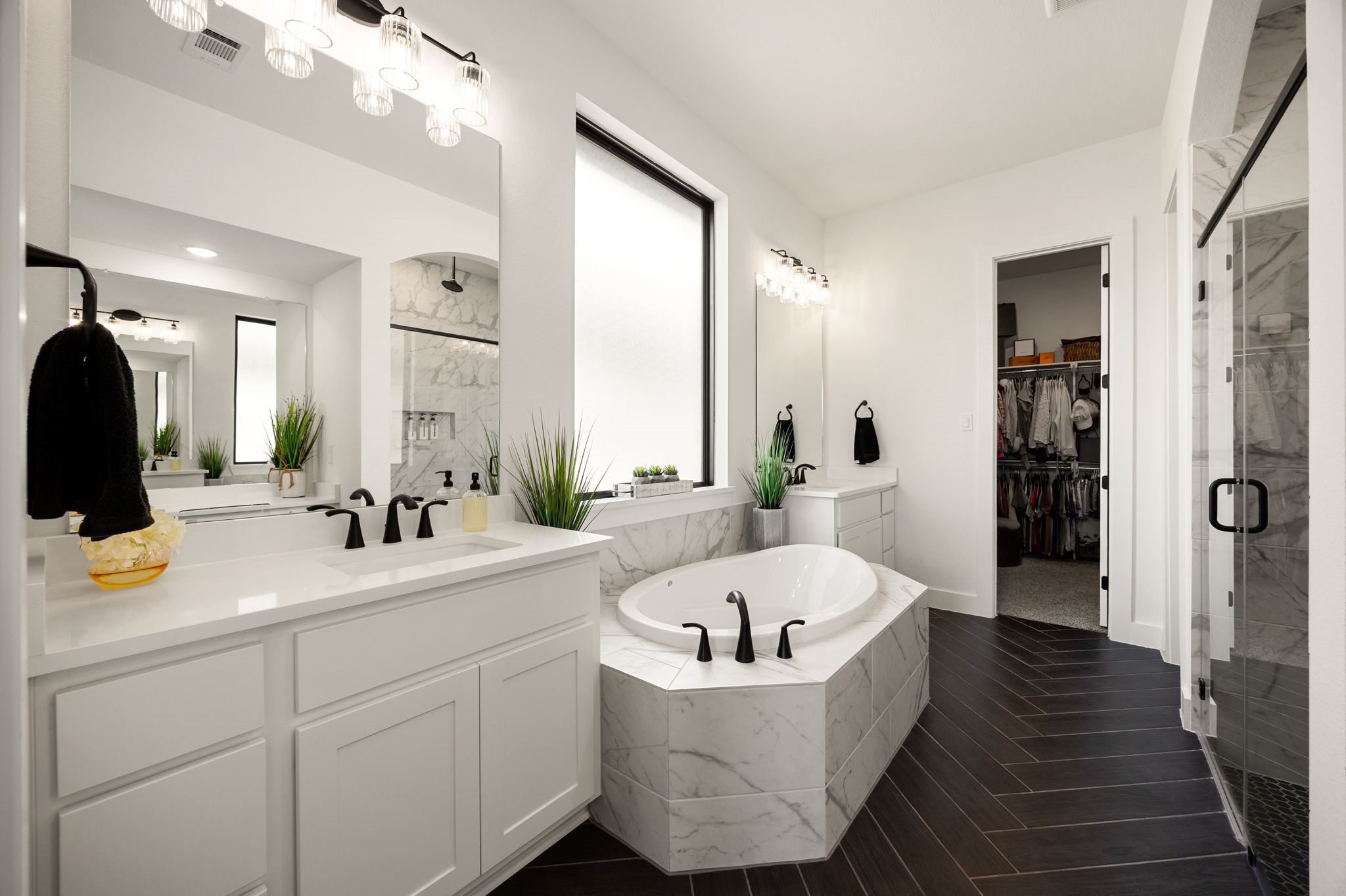 A spa-like primary en-suite. This luxurious bathroom features a deep soaking tub surrounded by marble tile, a large frosted window for natural light, white vanities with quartz countertops, sleek matte black fixtures, rich dark herringbone tile flooring, and a seamless glass shower entry.