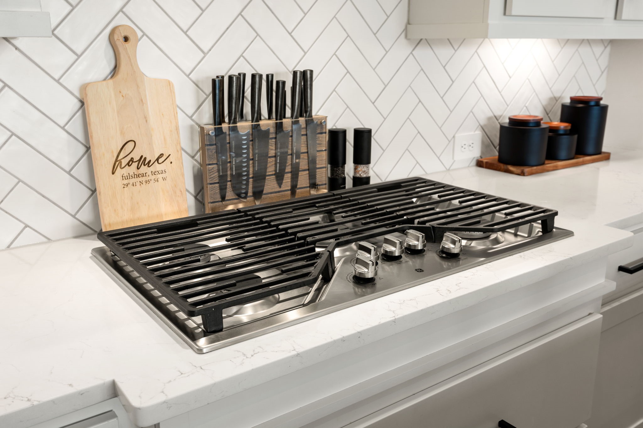 Premium culinary details. This chef-inspired cooking station showcases a high-end stainless steel gas cooktop with heavy-duty cast-iron grates, nestled into pristine white quartz countertops and framed by a classic white herringbone tile backsplash.
