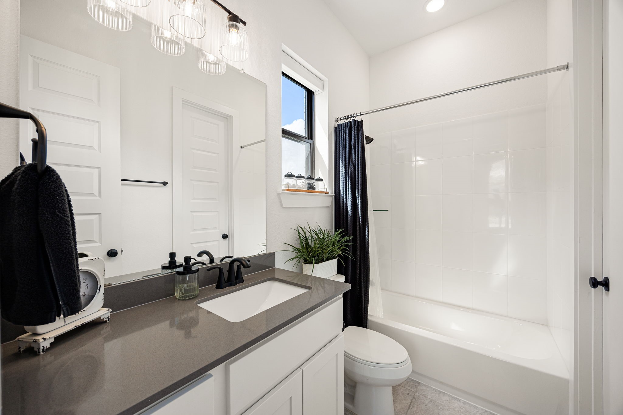 Immaculate and modern secondary bath. Crisp white cabinets contrast beautifully with premium grey countertops, an undermount sink, matte black hardware, custom vanity lighting, and a clean tub-and-shower combo.