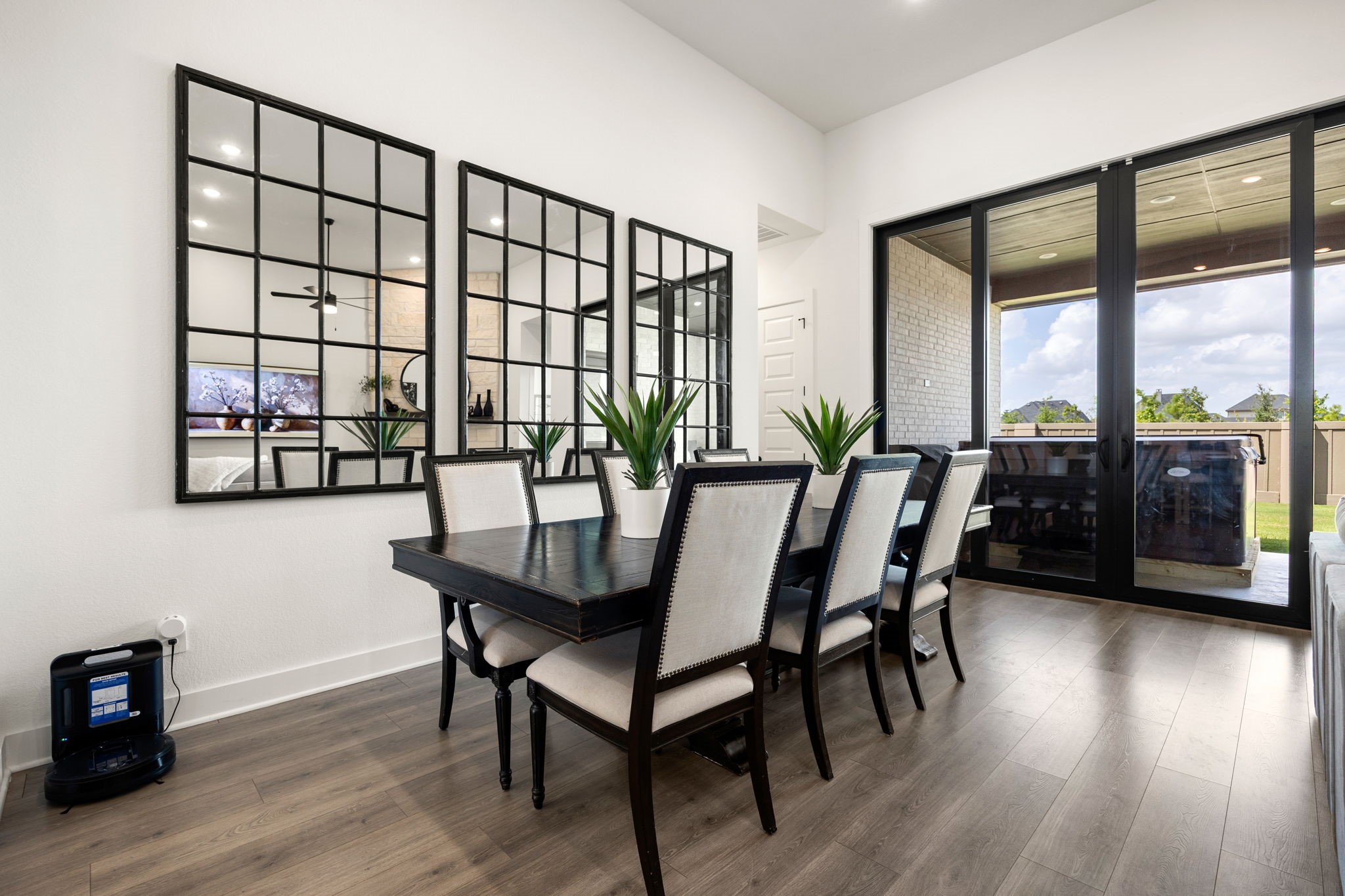 Bright and airy dining experience. Expansive wall-to-wall black glass sliders flood the room with natural light and lead out to a covered patio, beautifully showcasing the home's high-end wide-plank flooring, crisp white walls, and towering ceilings.