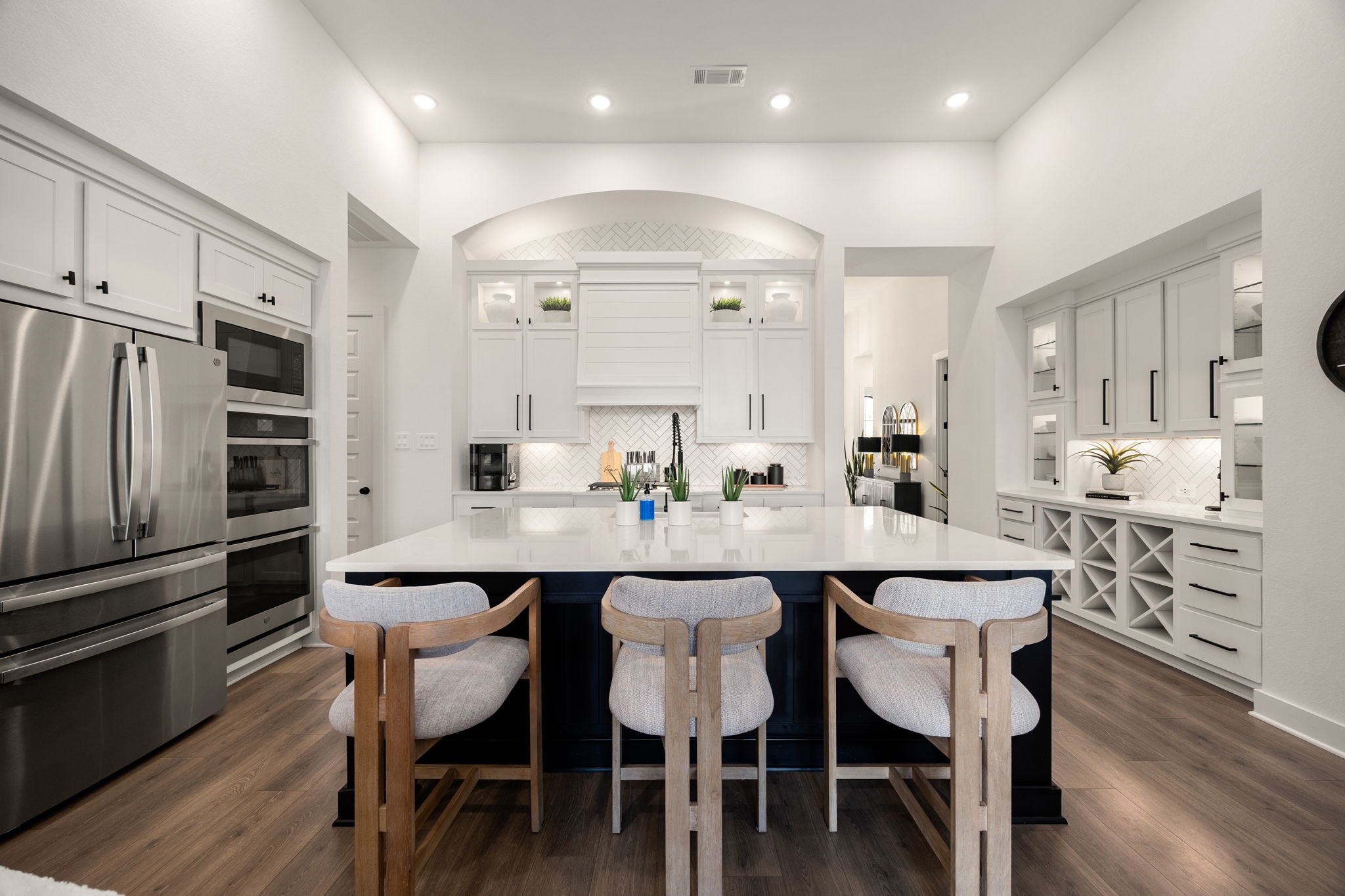 Gourmet cooking meets immaculate storage. This expansive view highlights high-end integrated stainless steel appliances, a massive quartz-topped island, extensive floor-to-ceiling white cabinetry, a built-in wine rack, and high-end wide-plank flooring under a grand architectural arch.