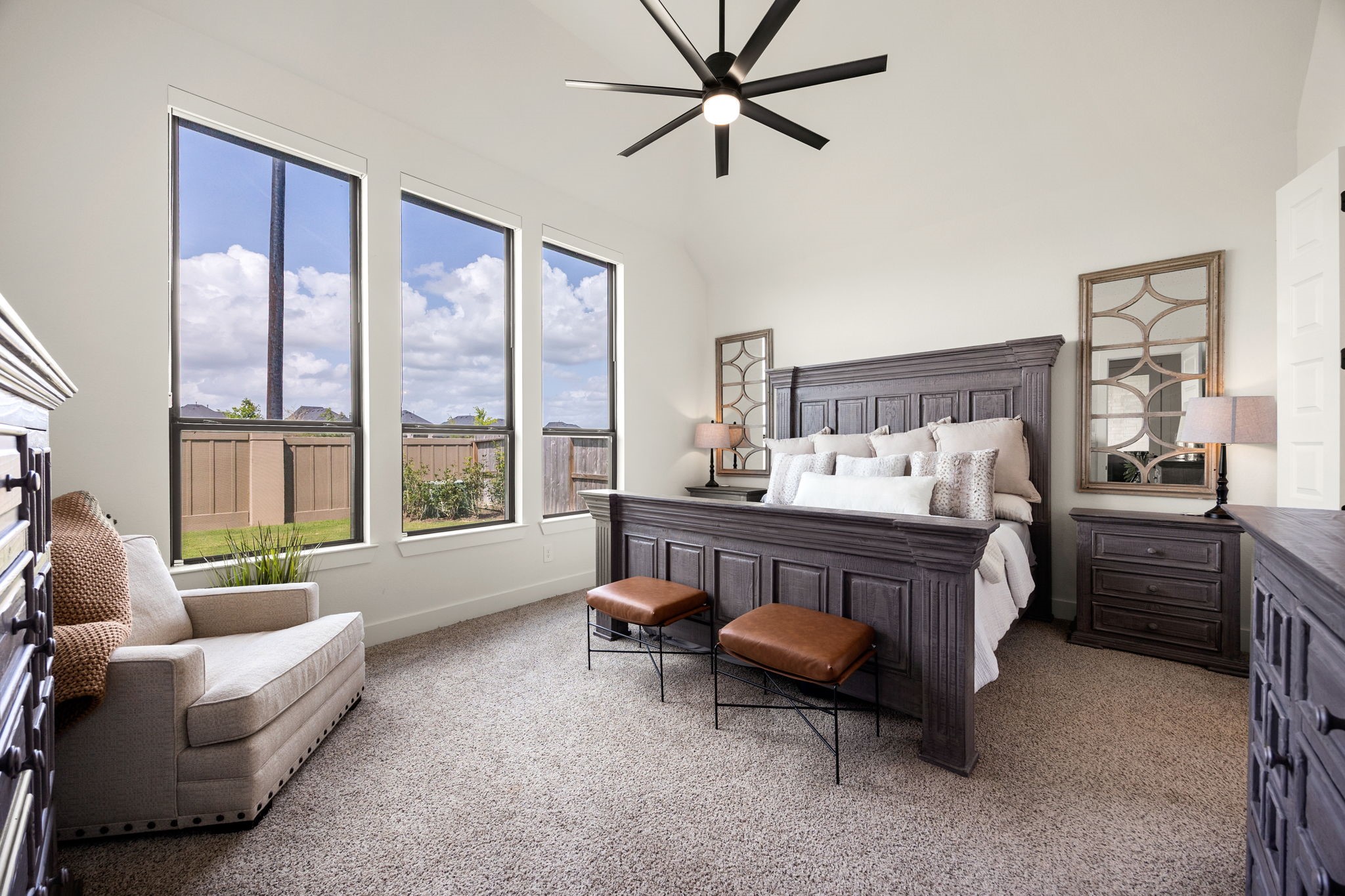 A serene primary retreat. Three towering, black-trimmed vertical windows with pull down blackout shades flood this spacious bedroom with natural light, showcasing plush carpeting, soaring vaulted ceilings, and a modern multi-blade ceiling fan with integrated lighting.