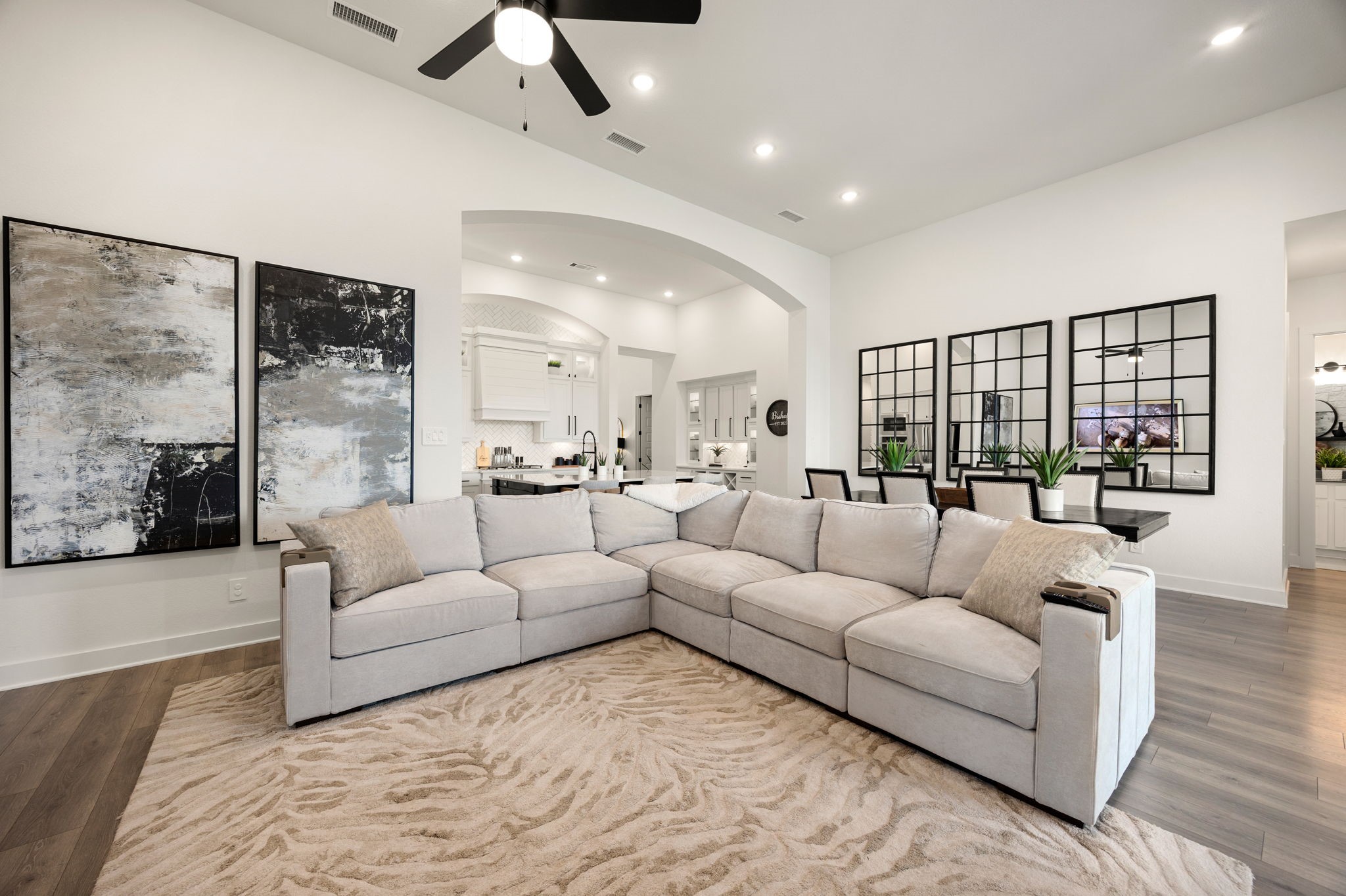 Designed for effortless modern living. This vast main room showcases stunning wide-plank flooring, crisp white walls, recessed lighting, and a grand arched opening that frames a bright view into the adjacent kitchen and dining spaces.