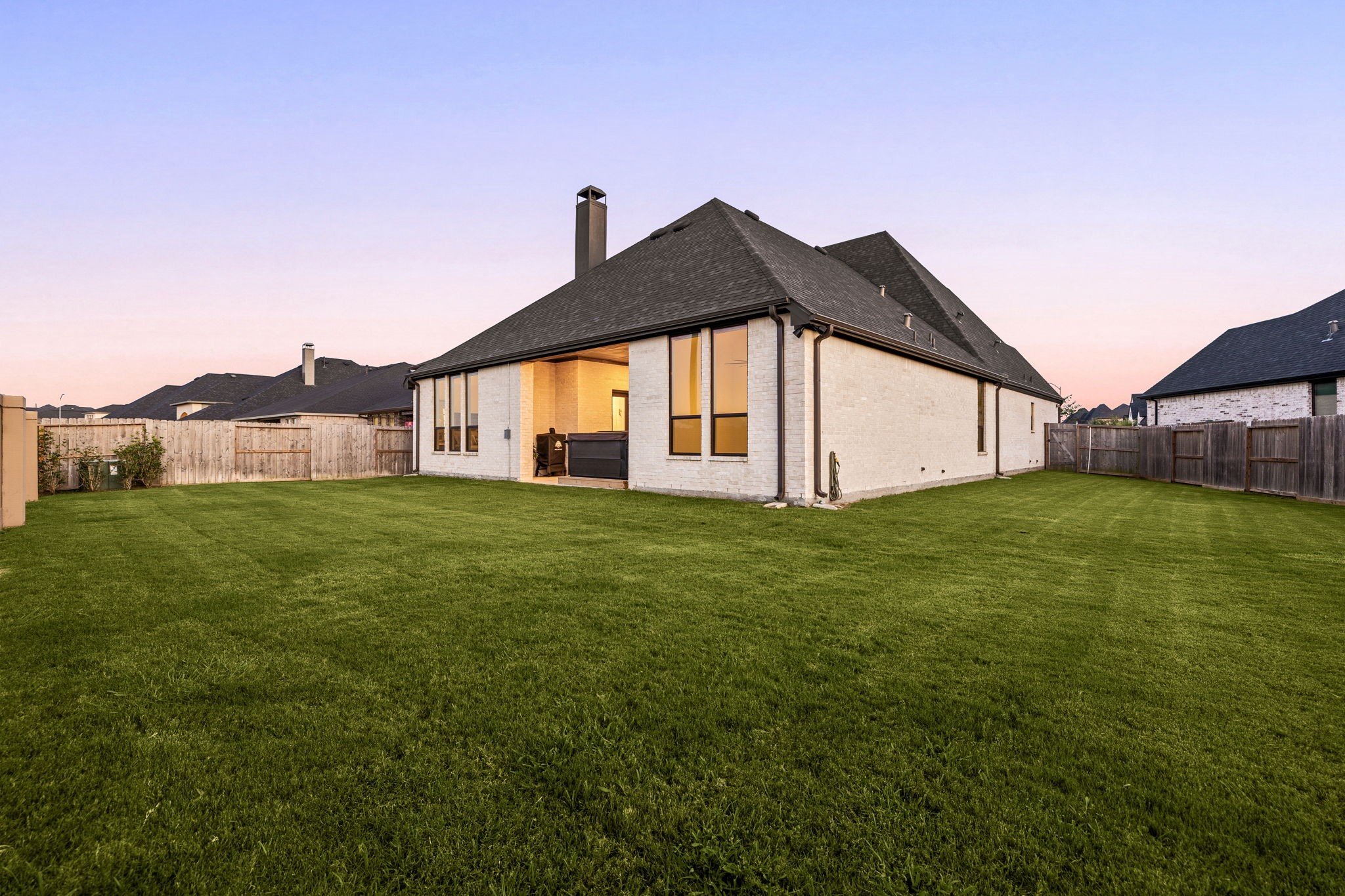An expansive, fully fenced backyard. A wide stretch of lush green lawn offers endless possibilities for outdoor activities, framed by a complete wooden privacy fence, a sleek brick chimney profile, and a covered back patio area.